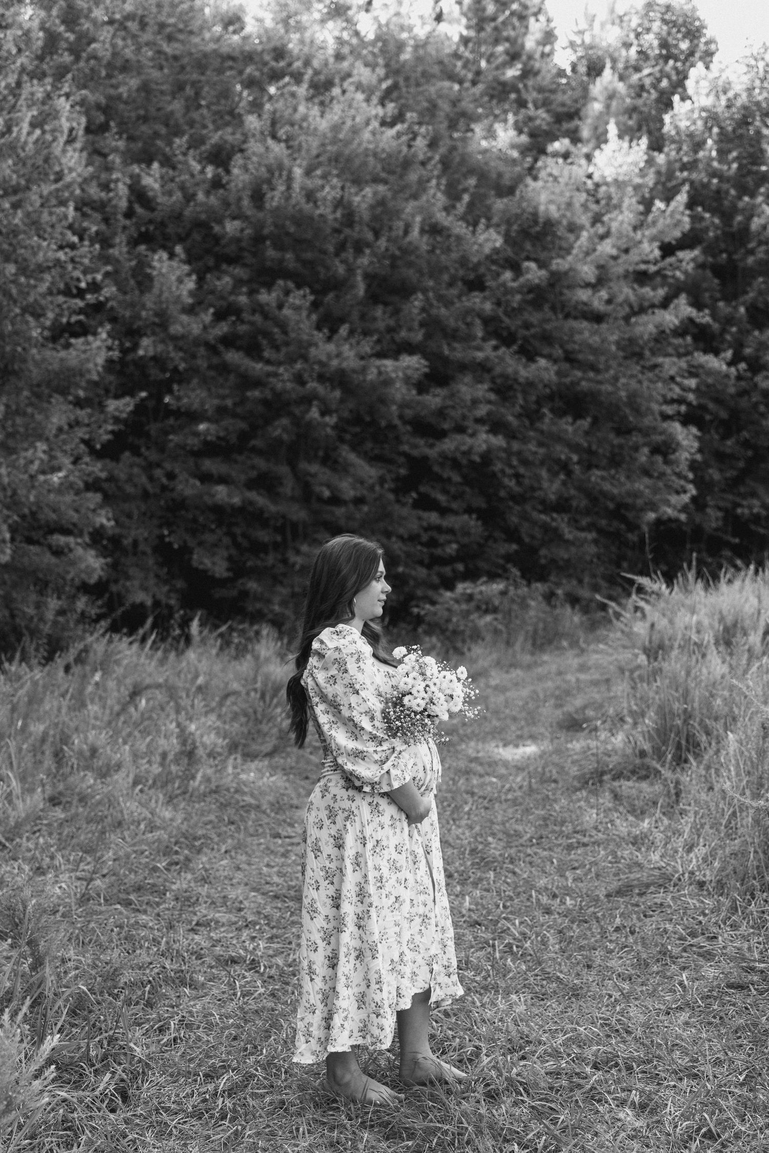 A figure in a floral dress stands in a grassy meadow holding wildflowers, with evergreen trees in the background.