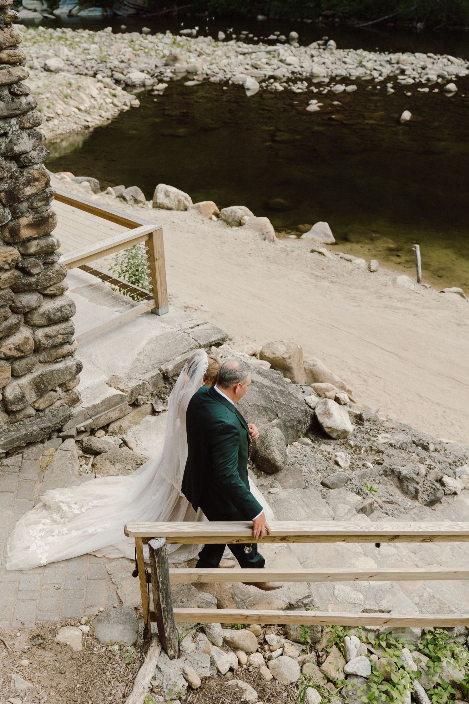 Bride and groom walk up wooden stairs near a rocky riverbank, her veil flowing in the breeze.