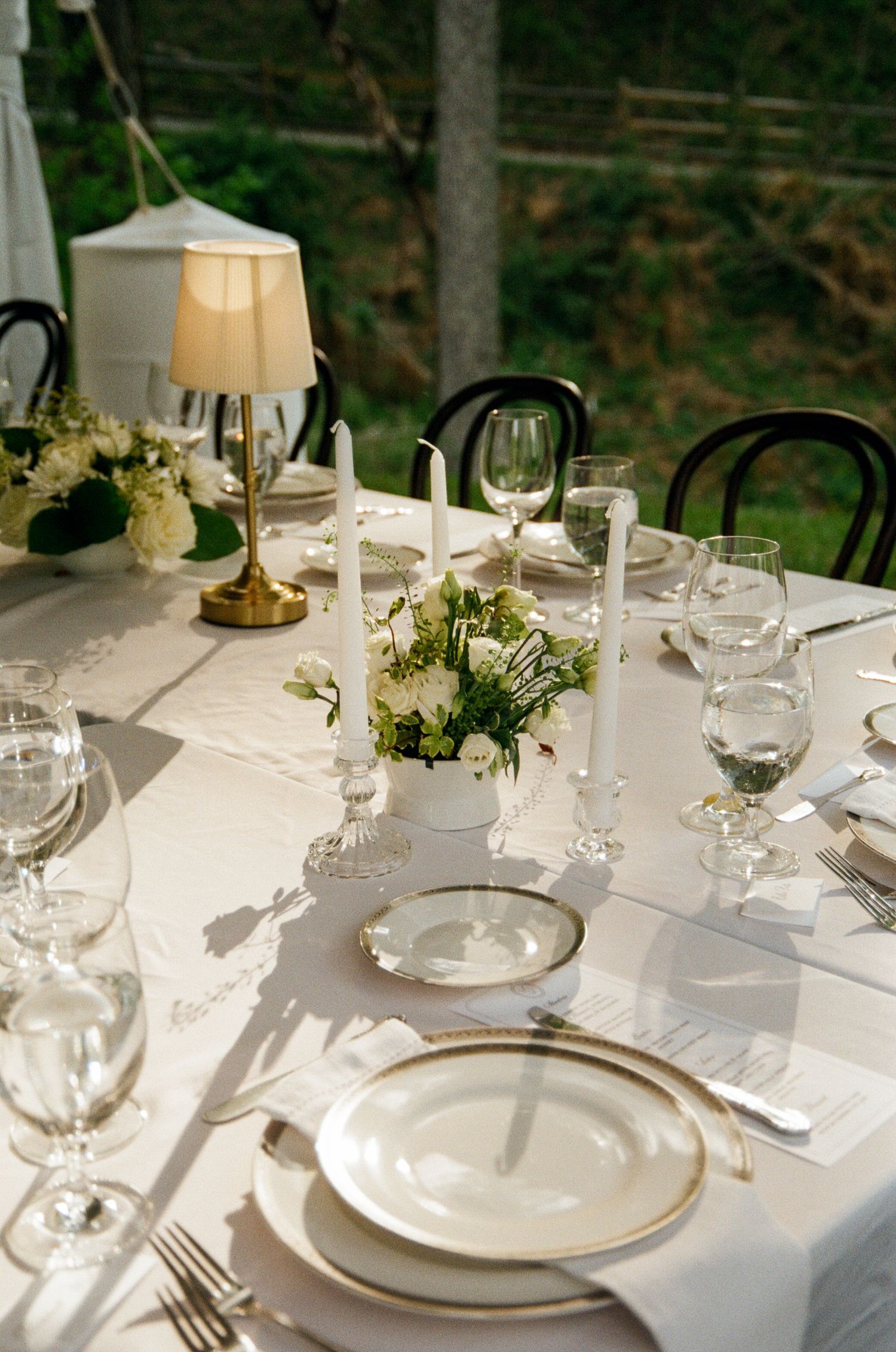 Elegant outdoor wedding table setting with white linens, candles, floral centerpiece, crystal glasses, and gold-rimmed plates.