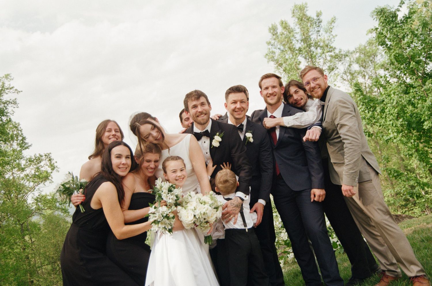 Wedding party posing outdoors, bride in white gown holding bouquet, groom in tuxedo, bridesmaids in black dresses.