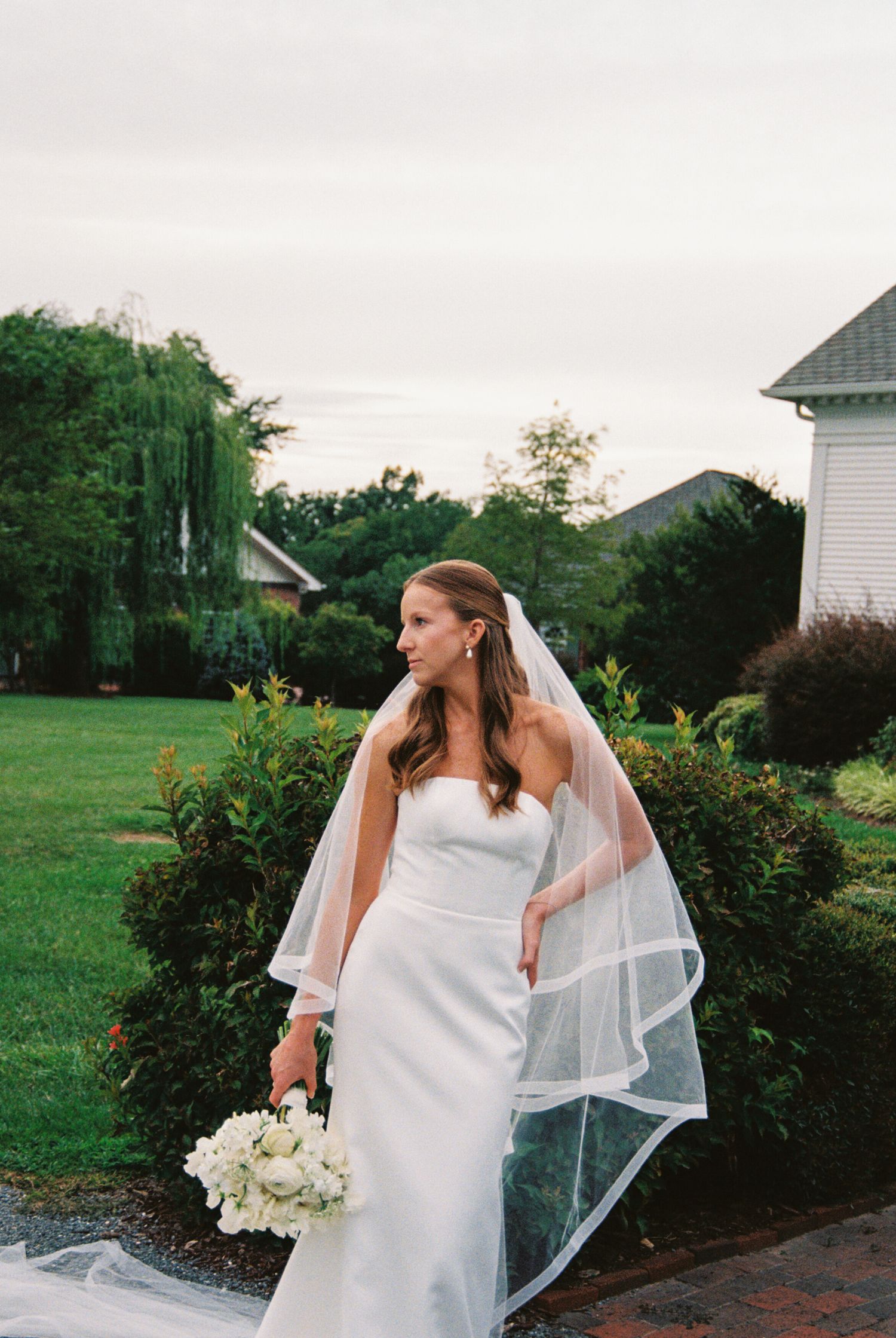 A bride in a sleek white wedding dress and flowing veil poses in a garden setting during sunset.