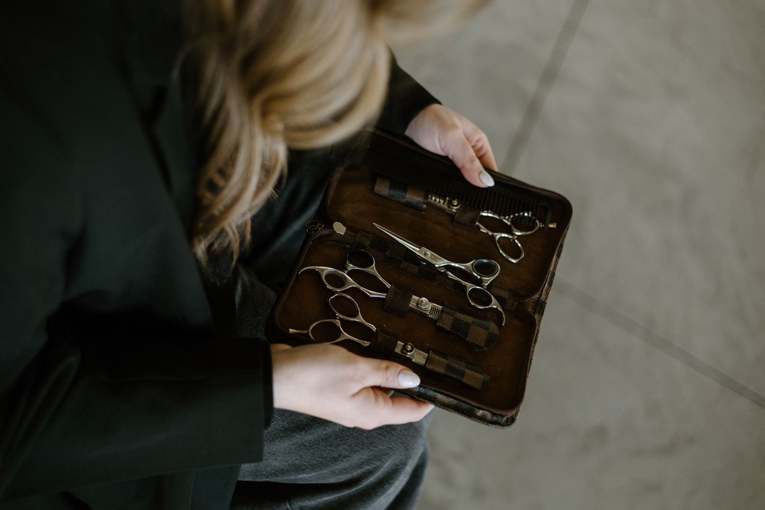 Professional hairstylist opens brown leather case containing various metal hair cutting shears and tools.