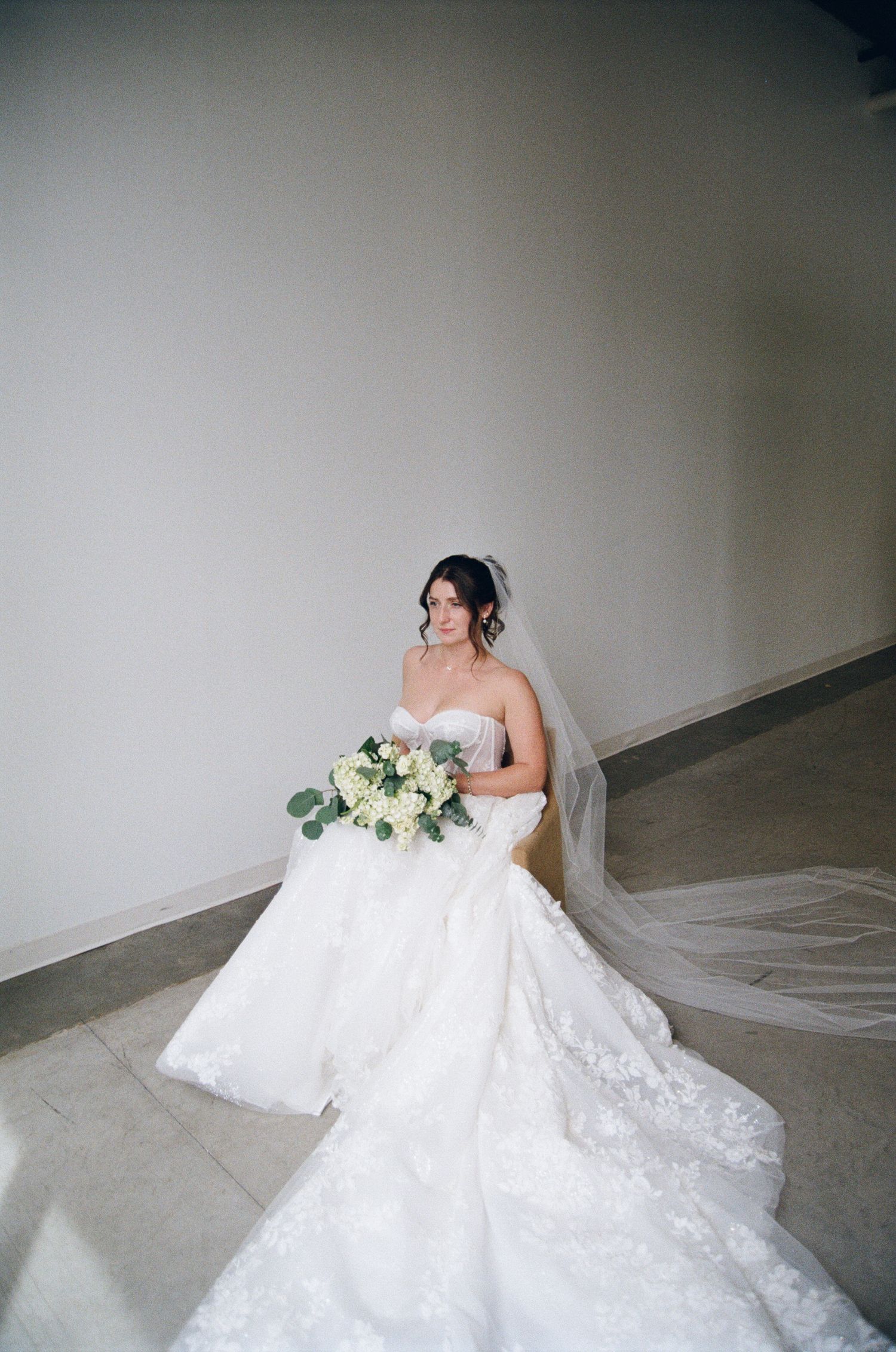 A bride in a stunning strapless white lace gown with a long train holds a delicate bouquet of white and green flowers.