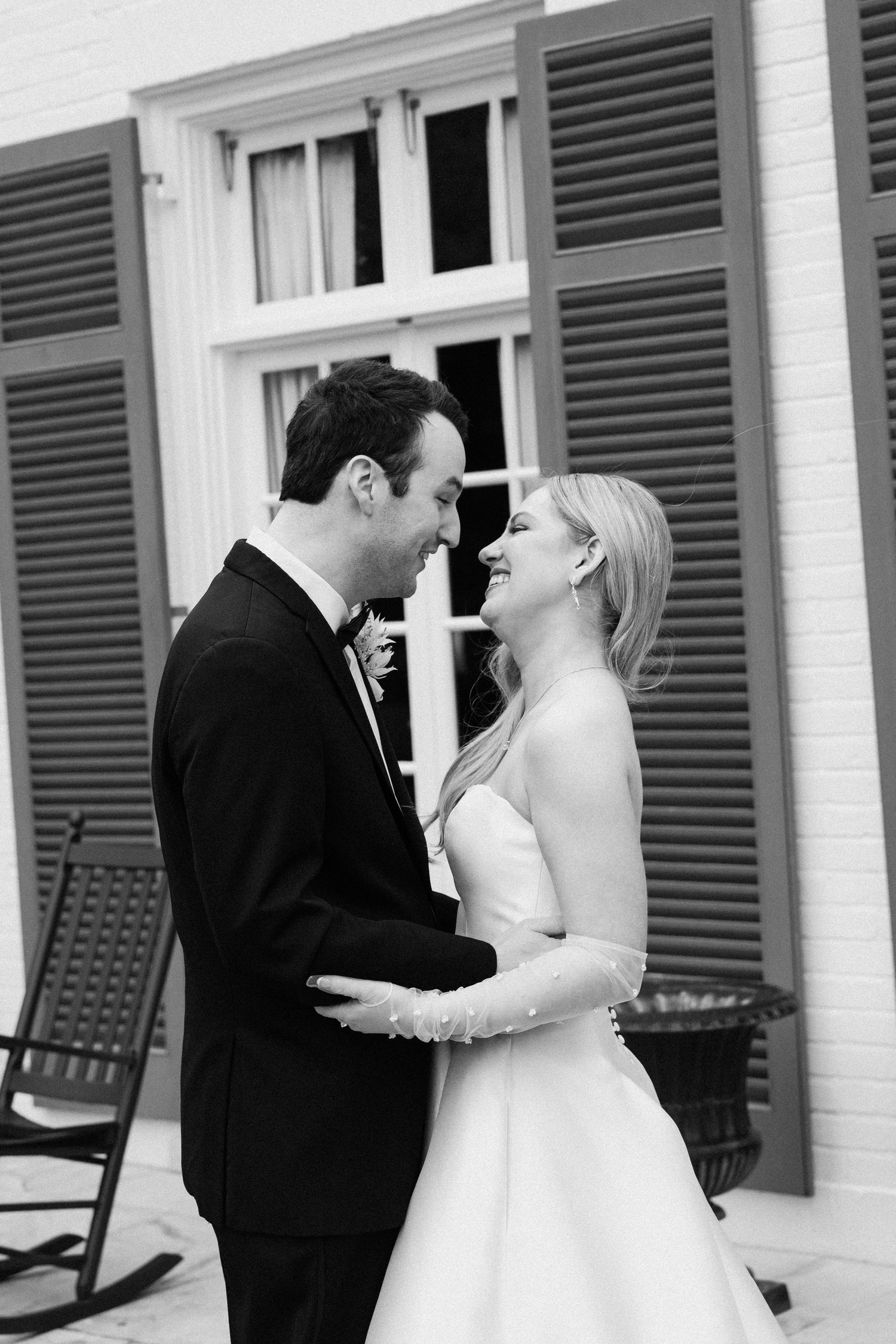 Black and white photo of a smiling bride and groom embracing outdoors near shuttered windows on their wedding day.