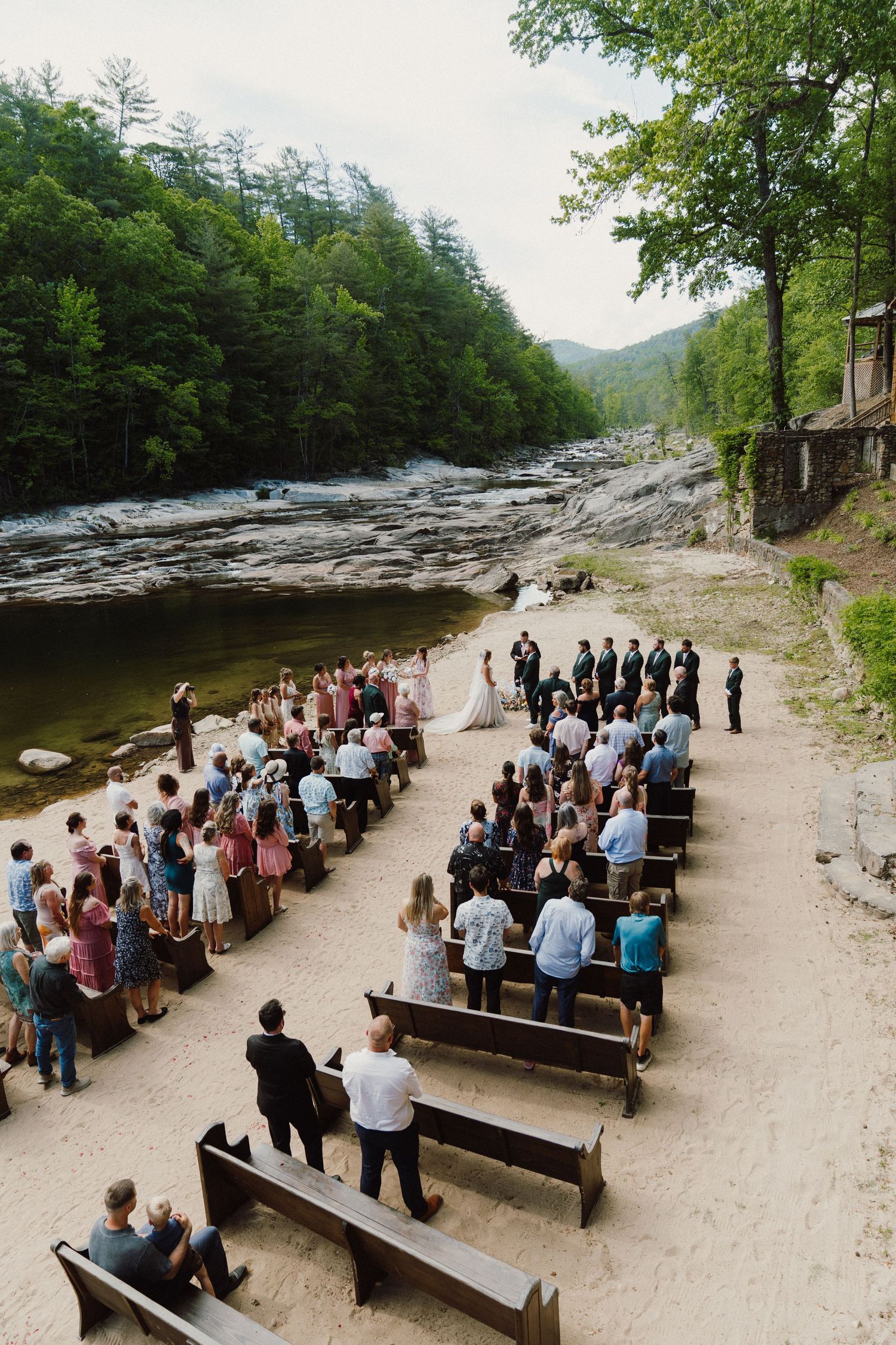 Outdoor wedding ceremony on sandy riverbank with wooden pews, lush forest backdrop, and mountains in distance.