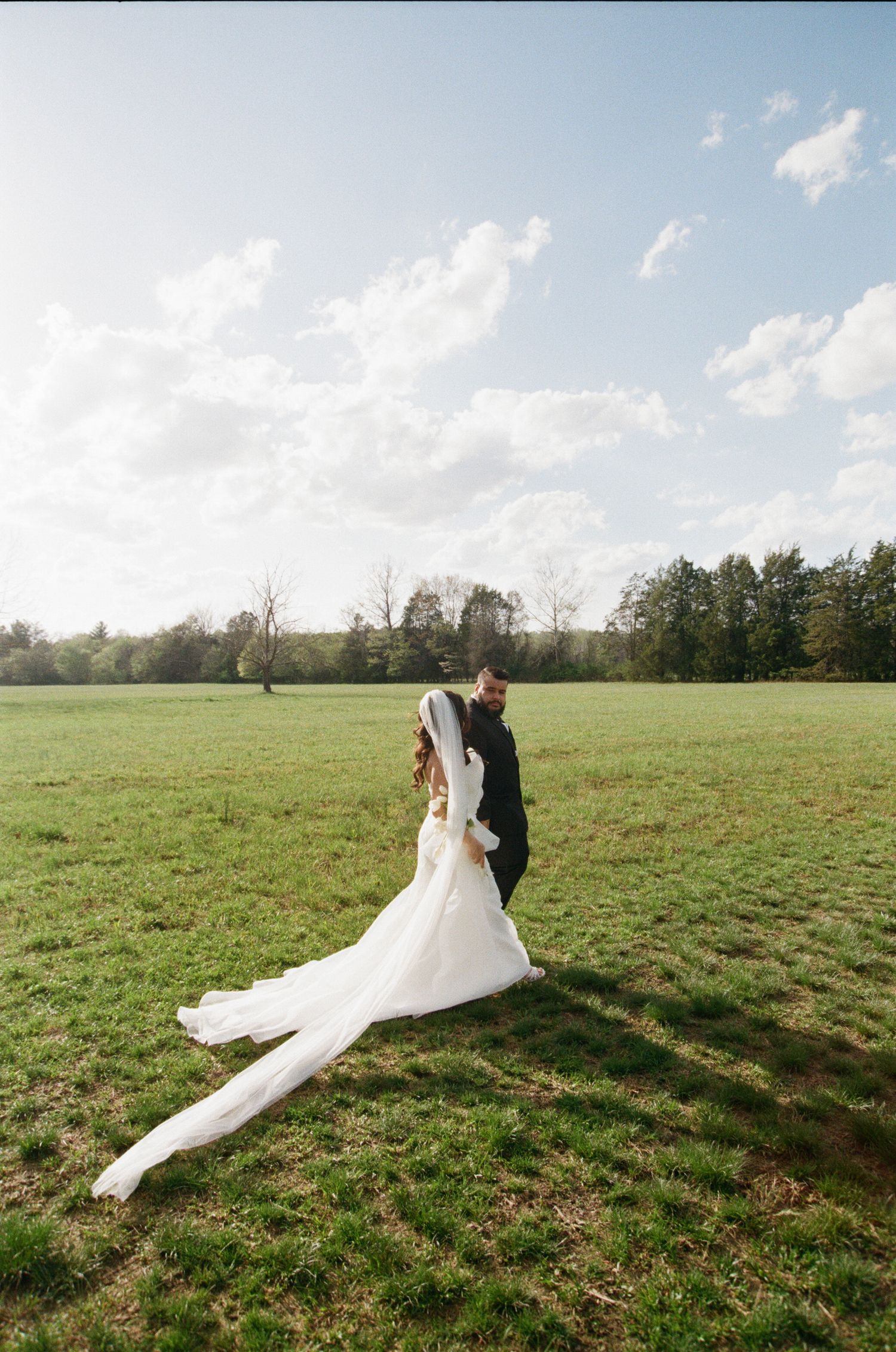 A couple walks together in a green meadow on their wedding day as the bride's white dress train flows behind them.