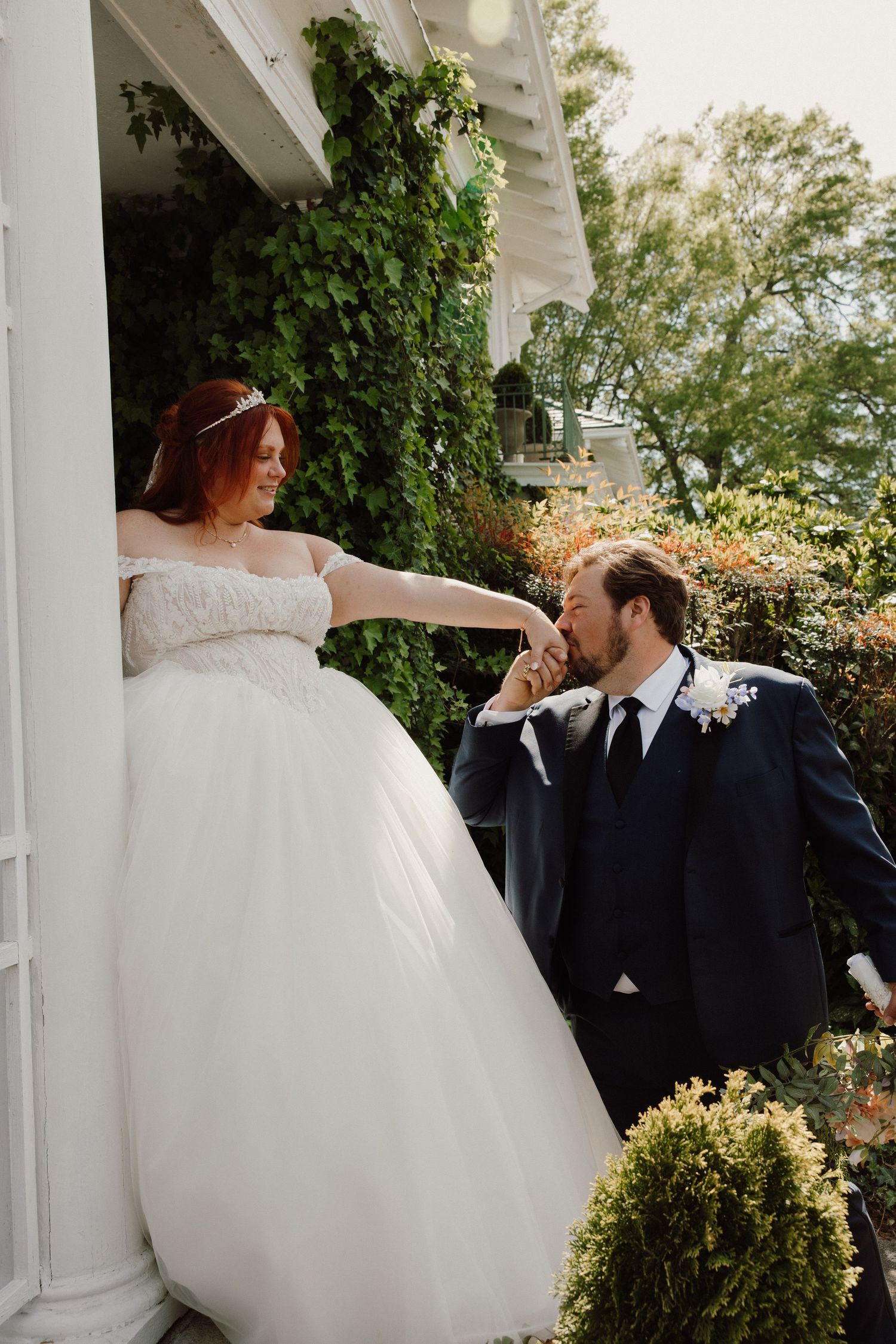 Groom in navy suit kisses bride's hand as she stands on porch in off-shoulder white ballgown at outdoor wedding.
