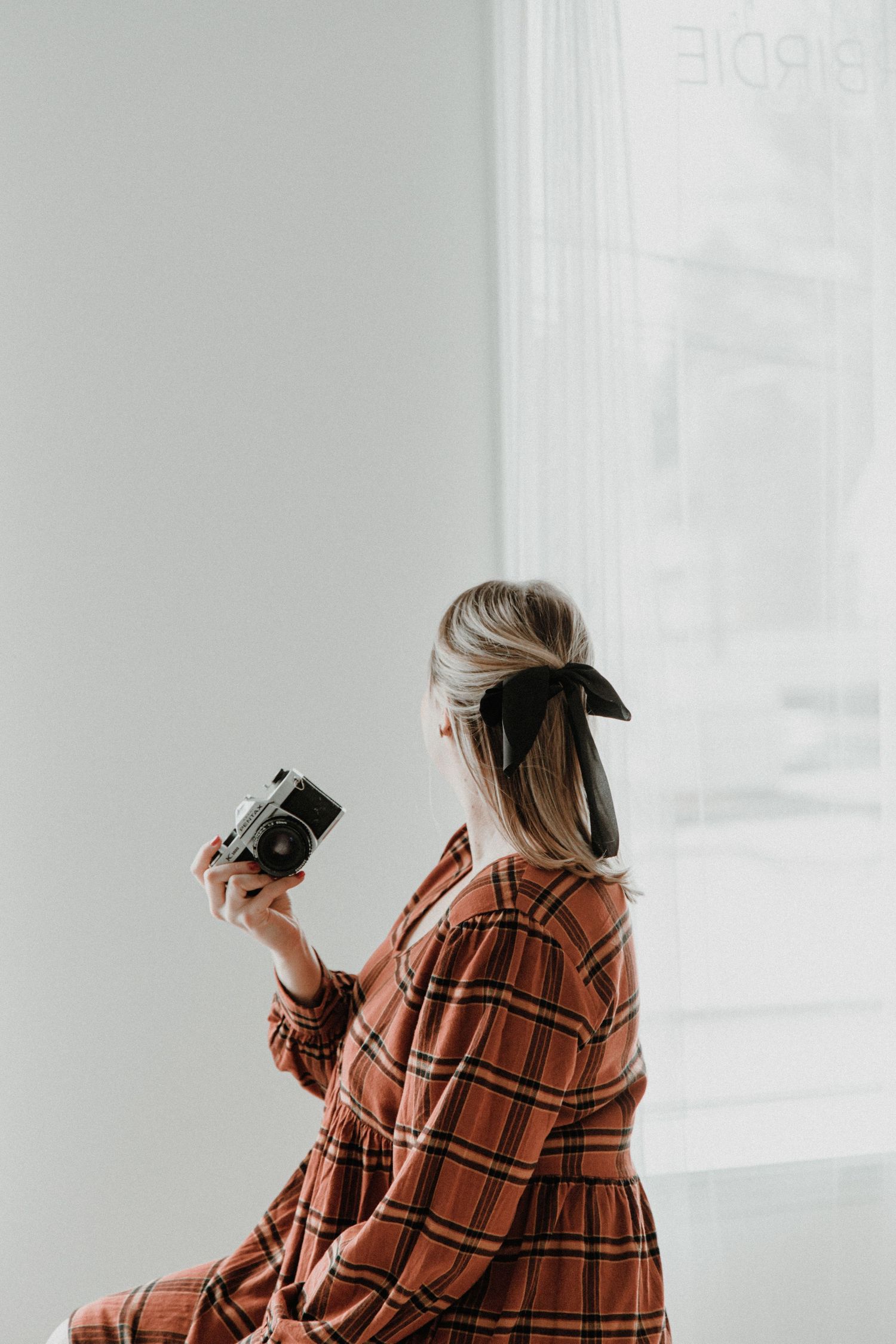 Person in plaid dress holding vintage camera near bright window with natural lighting.