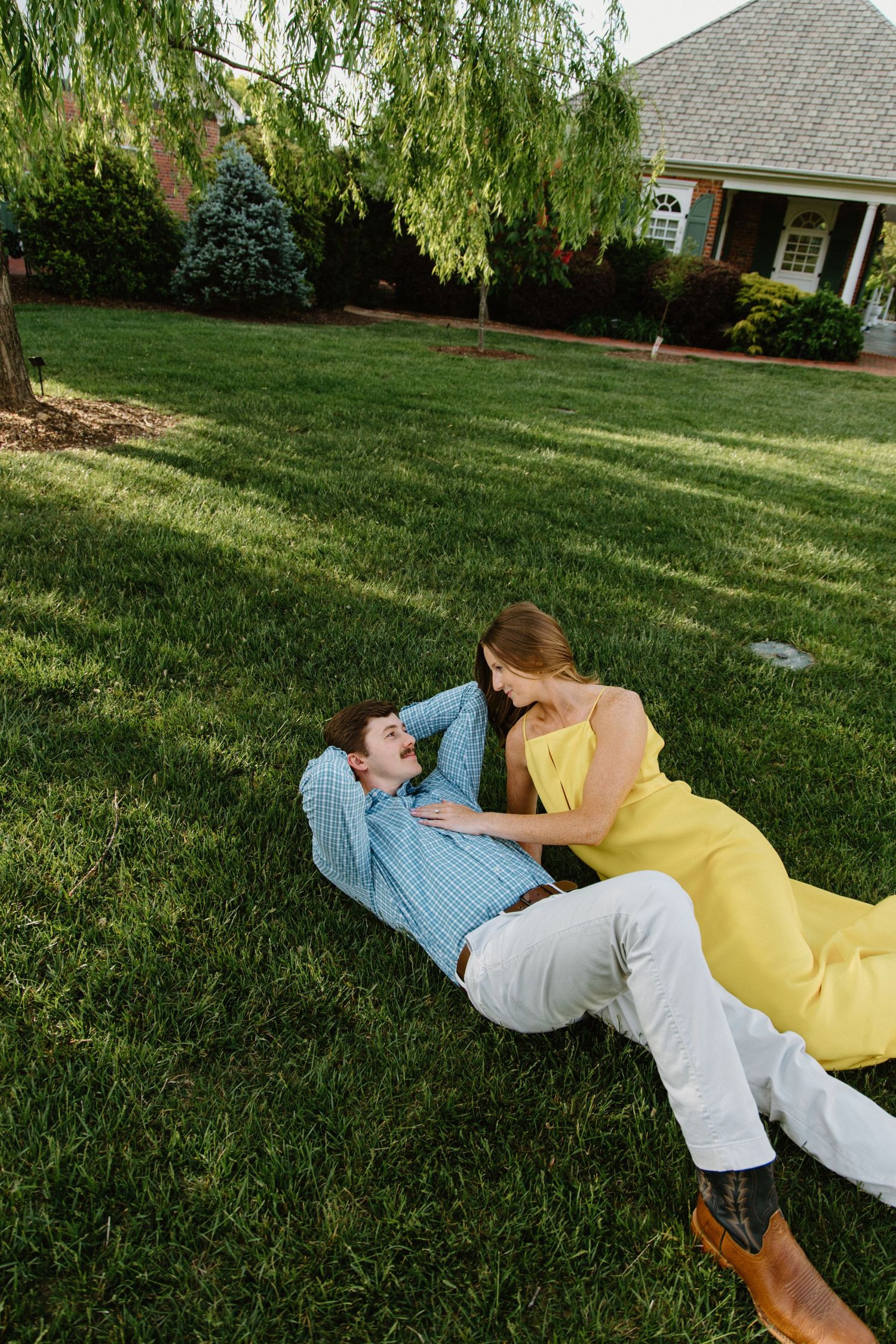 Two people lying on green grass having an intimate conversation in a backyard with a house in the background.