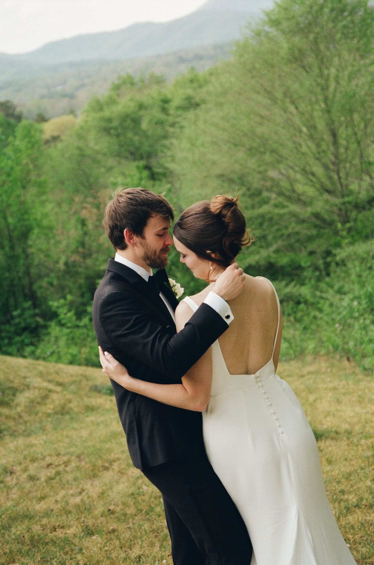 Bride and groom share a romantic embrace outdoors with lush green trees and mountains in the background.