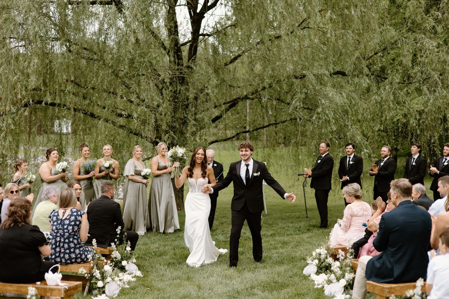 Outdoor wedding ceremony takes place under a weeping willow tree with guests seated on wooden chairs.