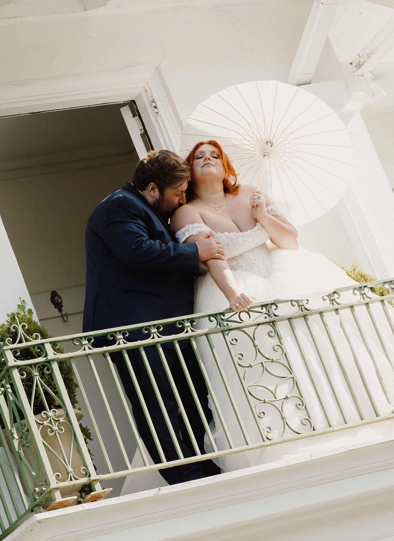 Bride in white gown holds parasol as groom in navy suit embraces her on elegant balcony with iron railing.