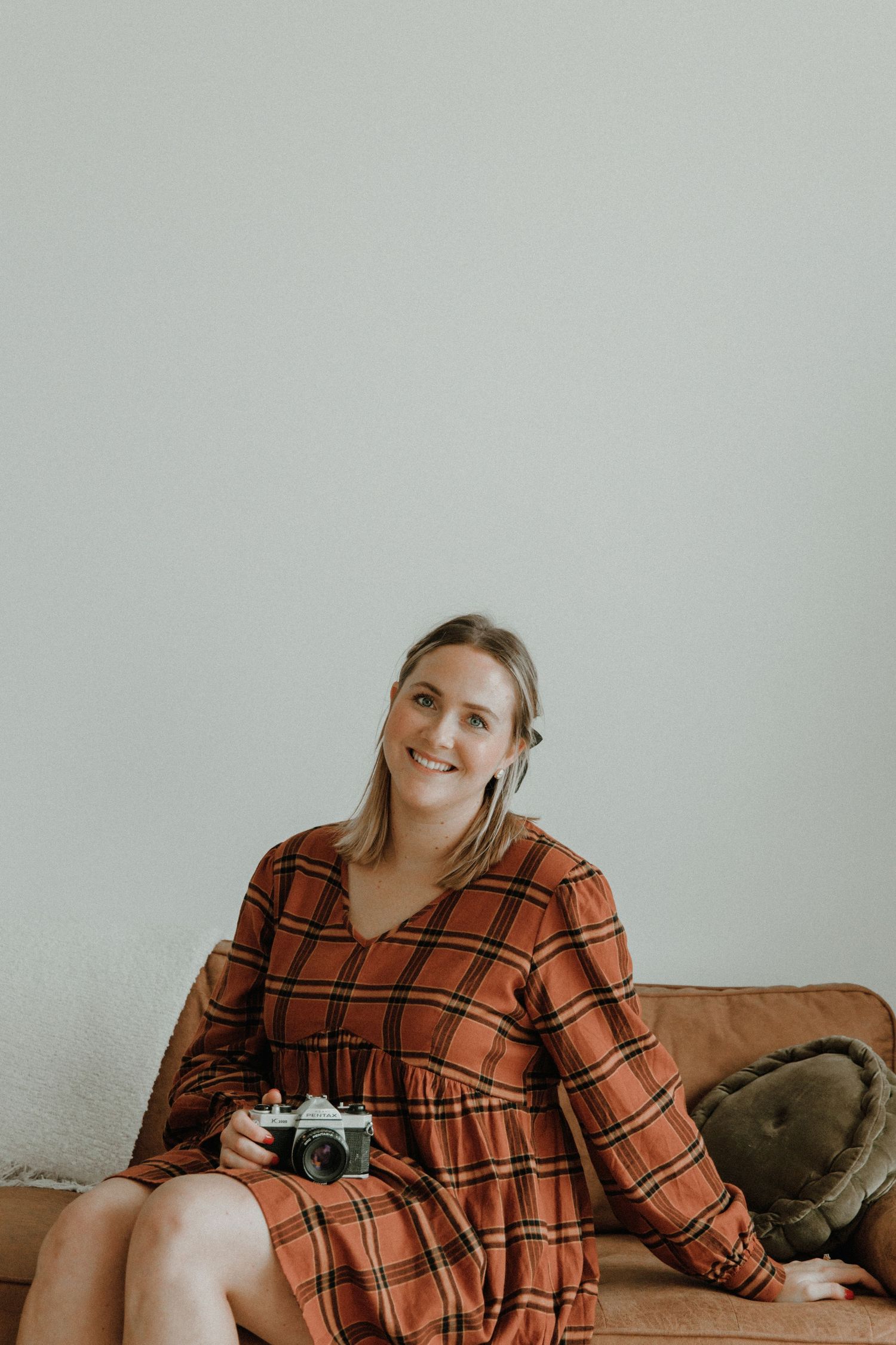 Person in orange plaid dress sits casually on couch holding a camera against light gray wall background.