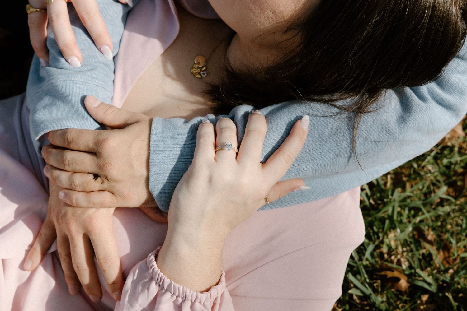 Two pairs of hands embrace tenderly in natural sunlight, one wearing pink and one wearing light blue clothing.