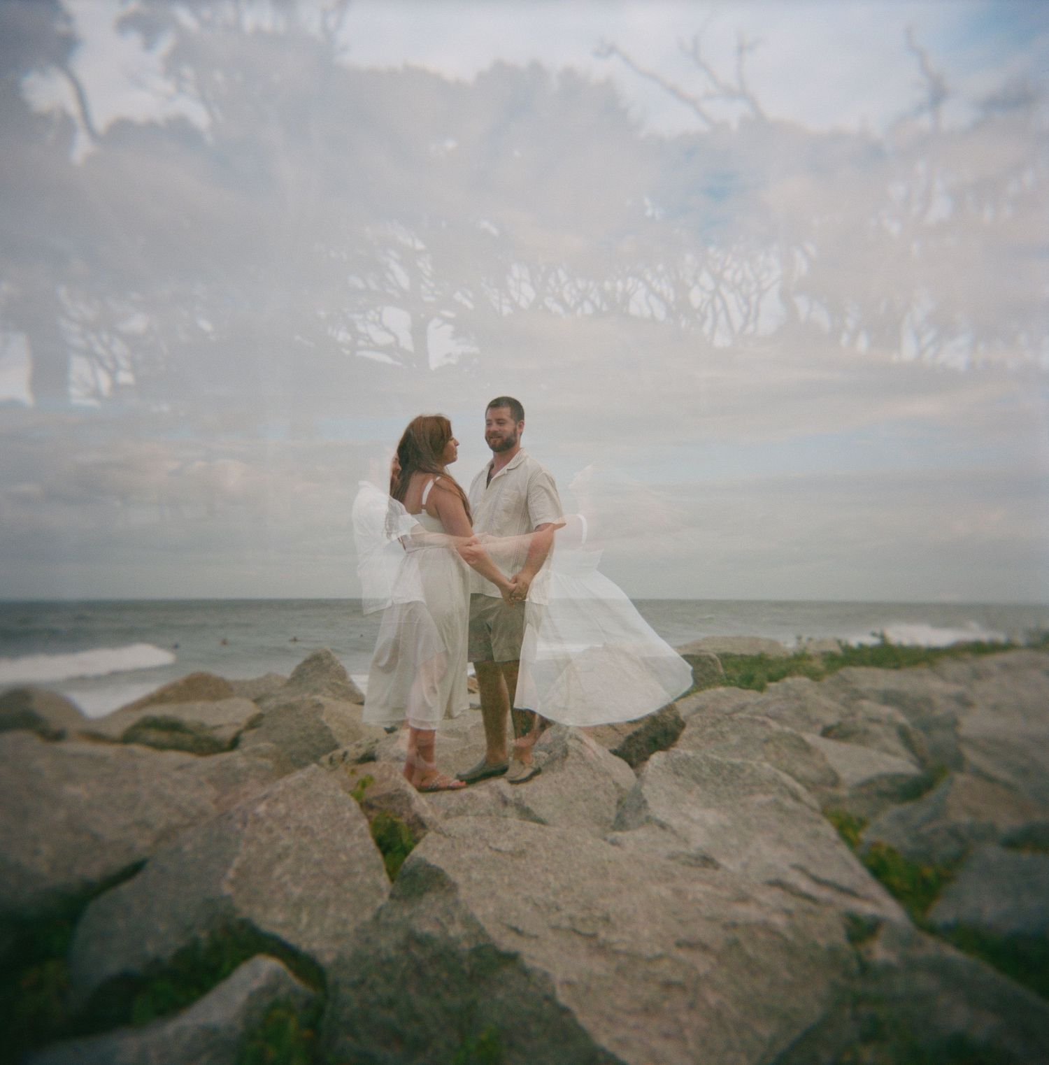 Couple sharing a romantic moment on coastal rocks as waves crash along the shoreline at sunset.