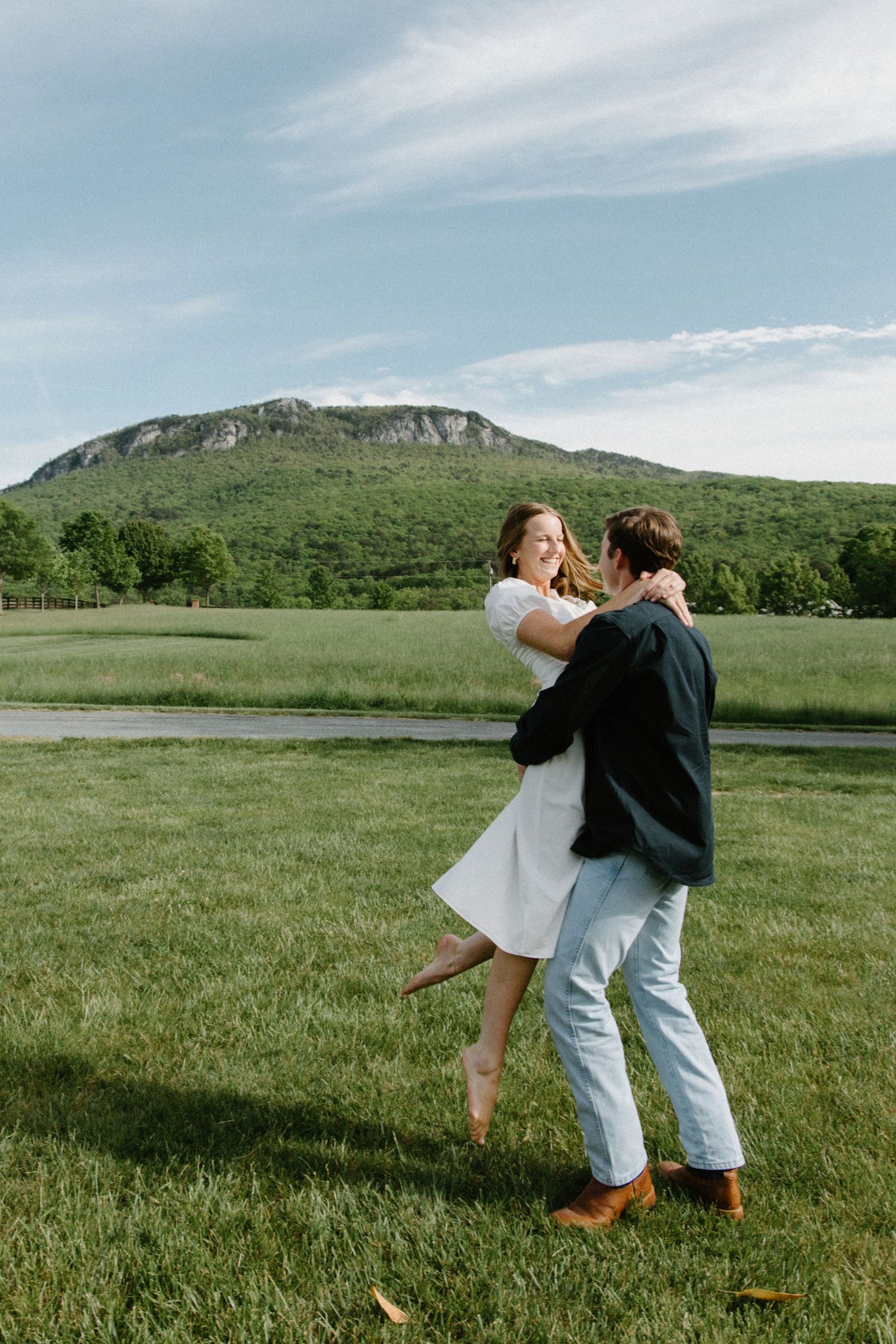 A couple dances playfully in a green meadow with mountains in the background on a sunny day.