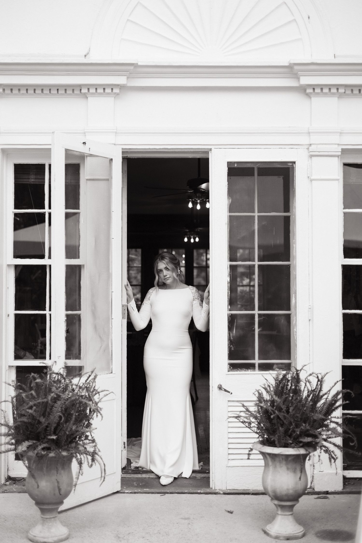 Elegant white wedding dress silhouette framed in french doors with ornate urns of greenery at entrance of classic building.
