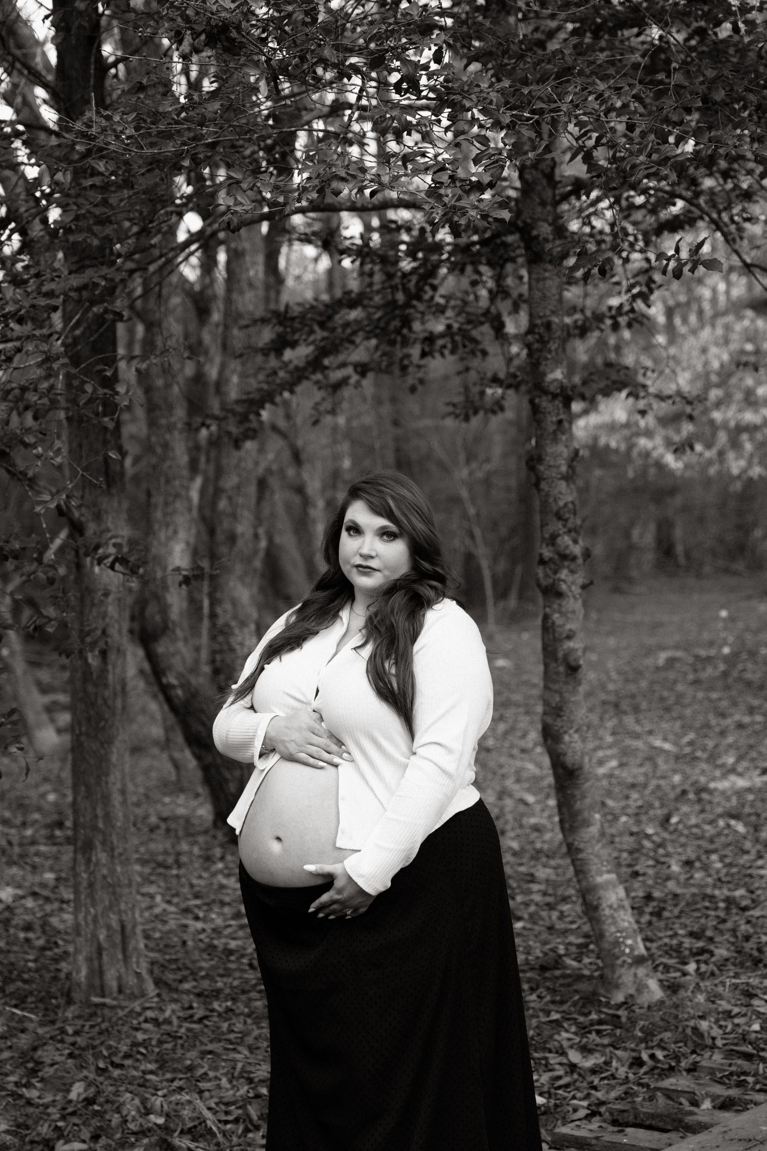 Black and white maternity portrait in a forest setting with woman in long skirt and white top cradling her baby bump.