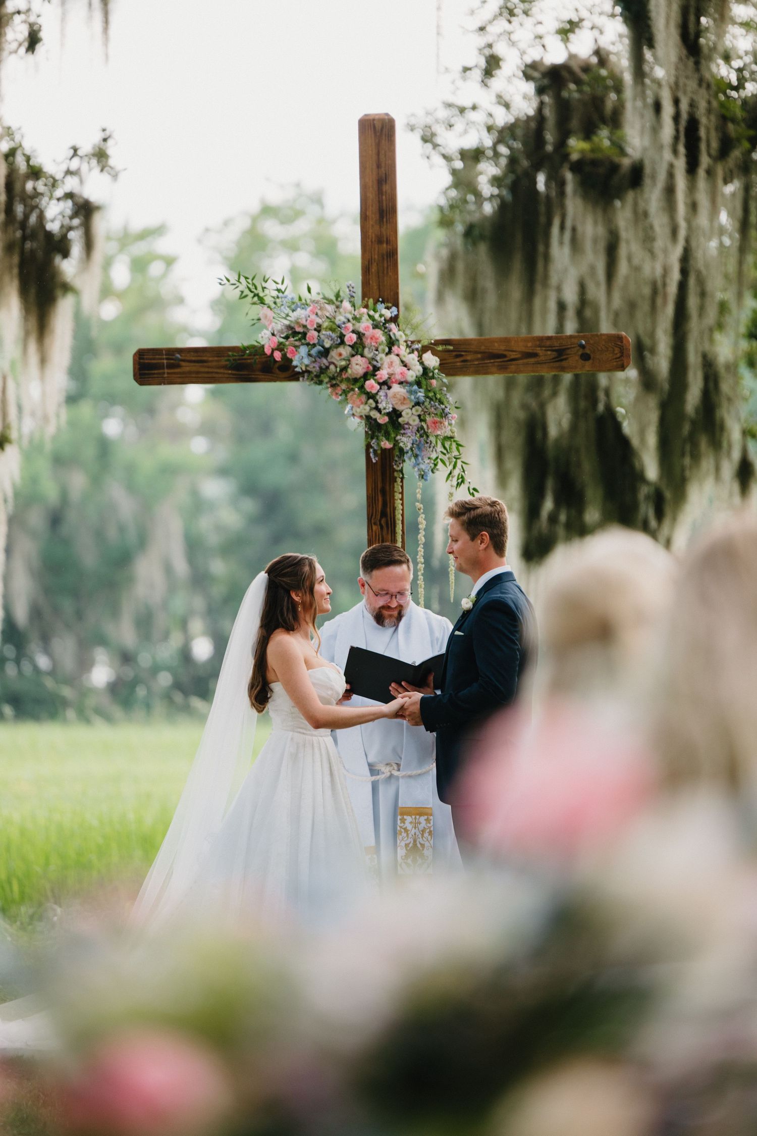 A wedding ceremony takes place outdoors beneath a wooden cross decorated with pink flowers and Spanish moss.