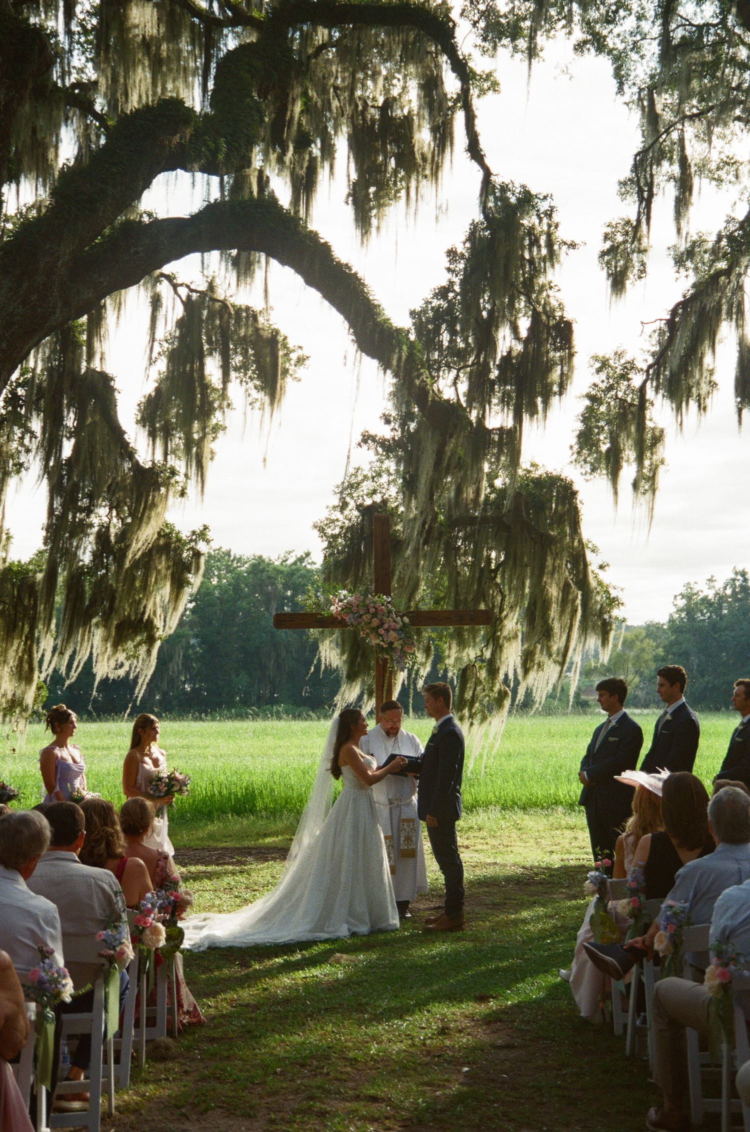 Wedding ceremony taking place under Spanish moss draped oak trees in a scenic outdoor setting at sunset.