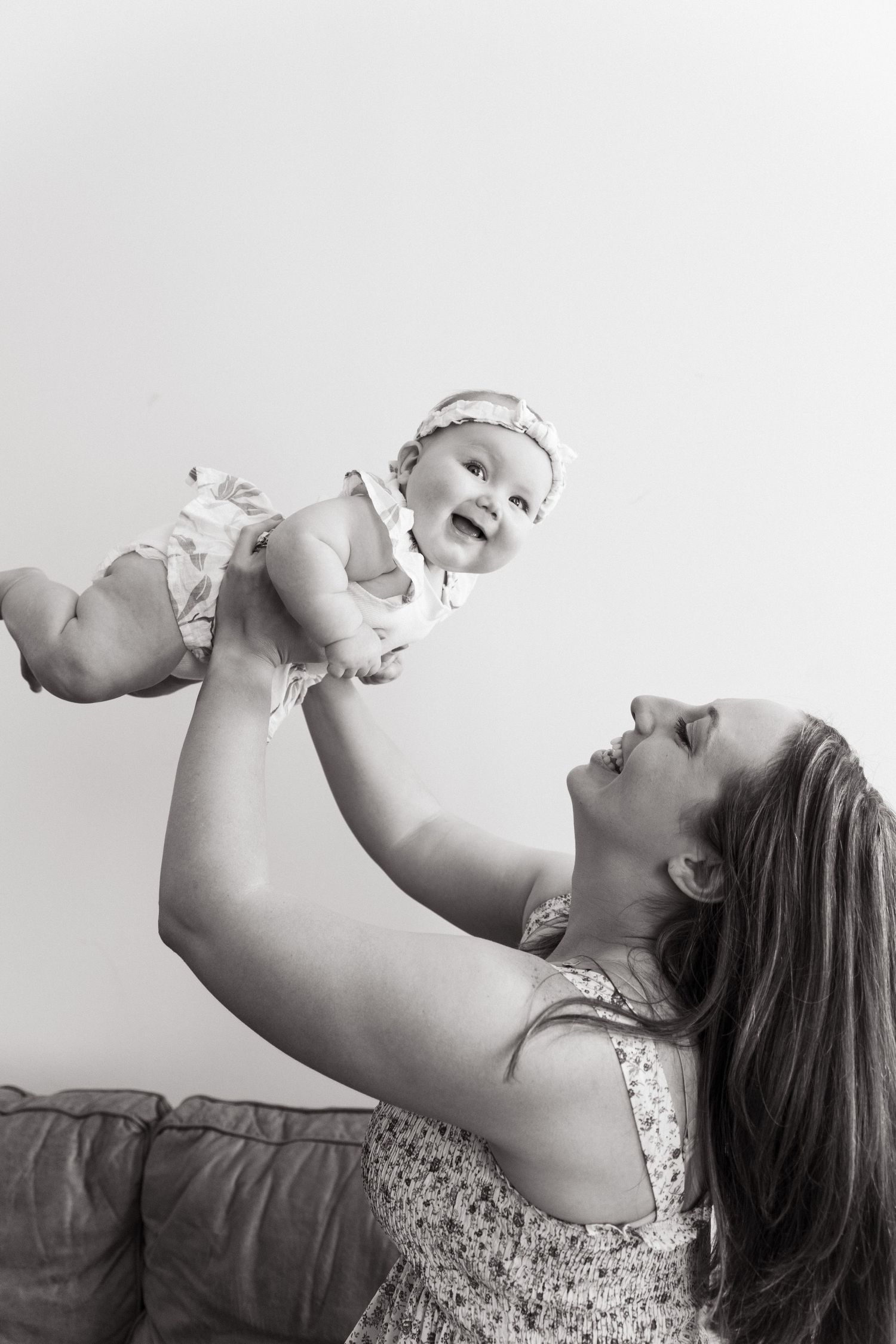 Mother in floral dress lifts laughing baby in black and white portrait.