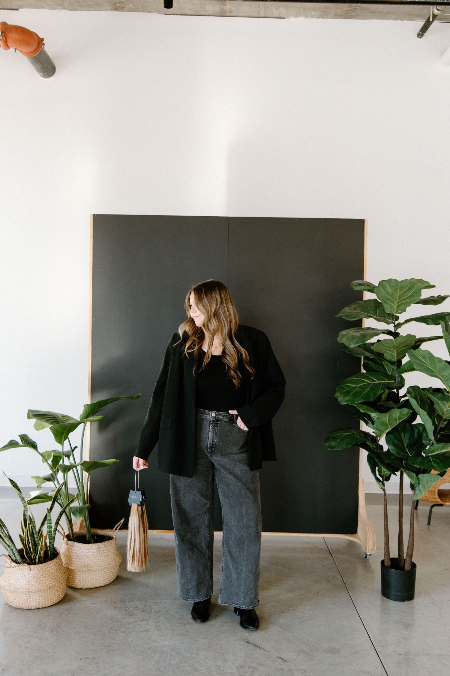 A person in a black blazer and wide-leg jeans stands against a dark backdrop with potted plants around them.