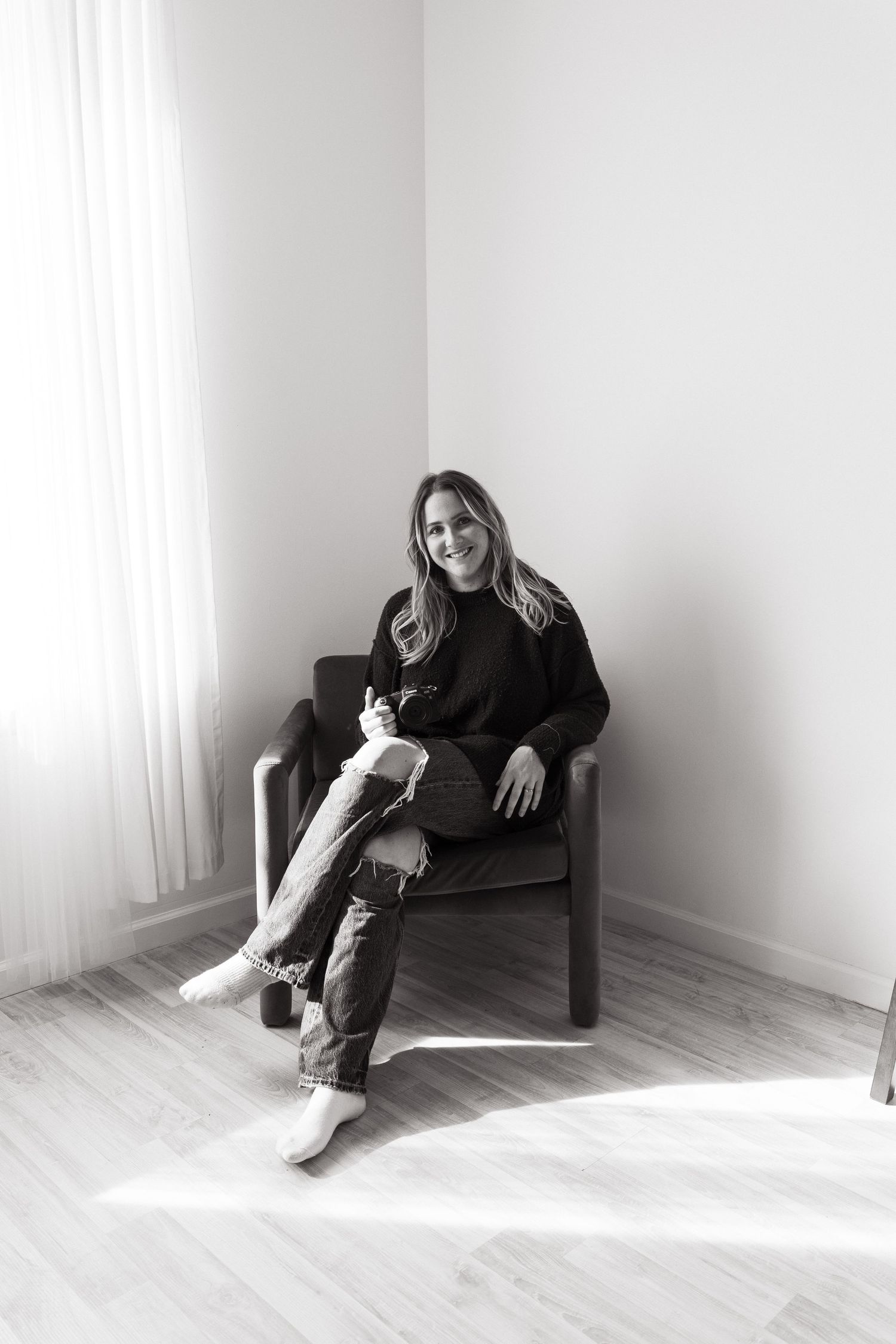 Black and white portrait of someone relaxed in an armchair near a bright window in a minimalist room setting.