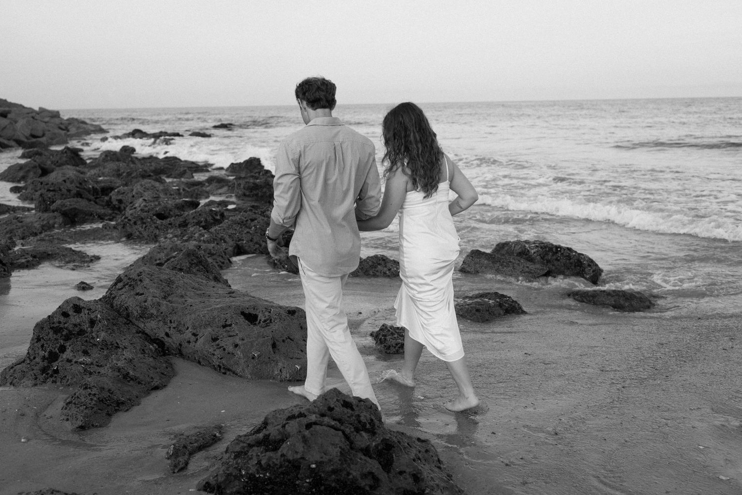 Couple in white outfits walking along a rocky beach shore during sunset against an ocean backdrop.