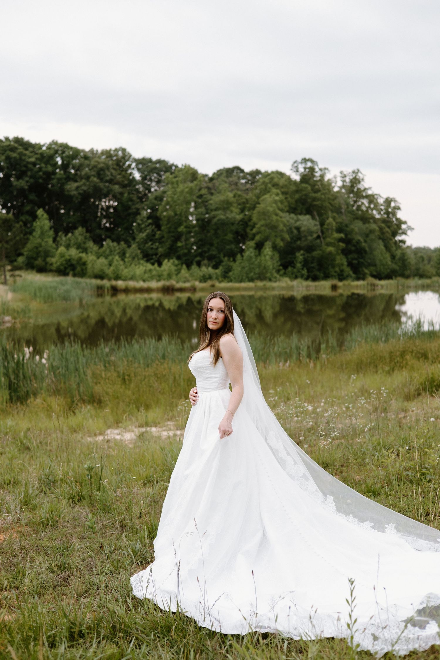 A bride in a flowing white wedding gown with long cathedral veil stands in a meadow beside a serene lake.