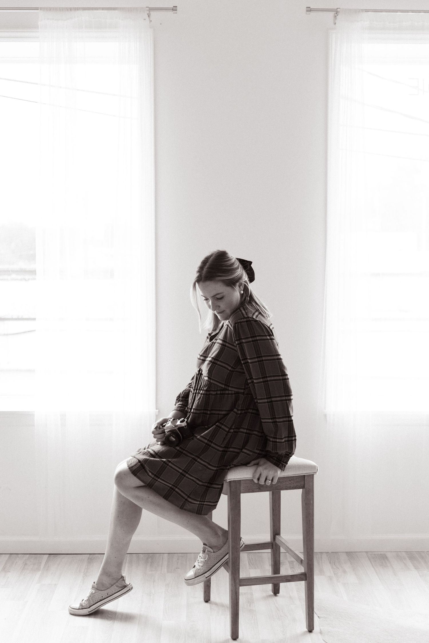 Black and white portrait of person sitting sideways on stool near bright windows.