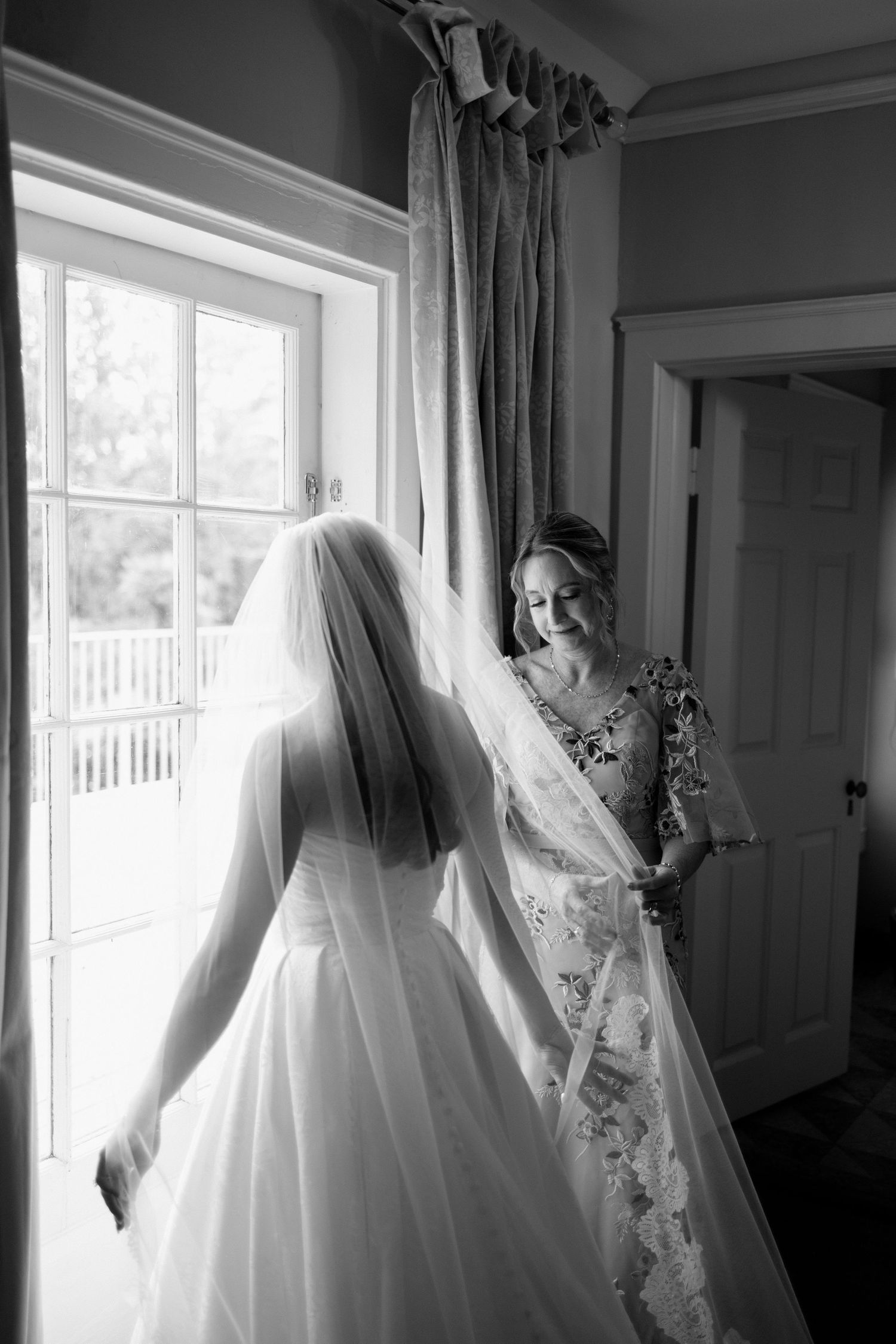 A wedding dress with a long veil hangs by a sunlit window in a bridal preparation room.