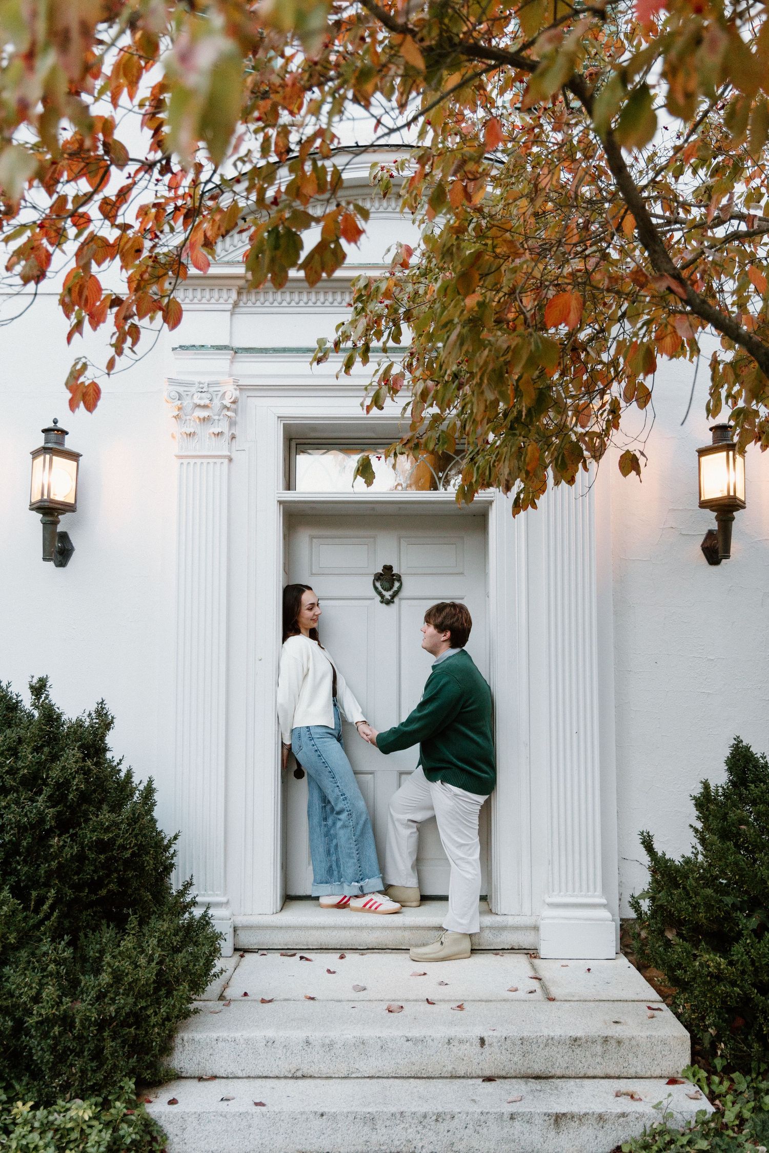 Couple sits on white columned porch steps surrounded by autumn leaves and hanging lanterns in romantic setting.