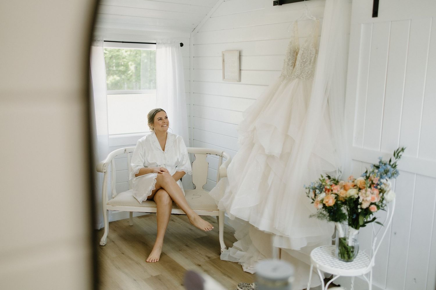 A white bridal robe hangs on display next to a chair and floral arrangement in a bright, airy dressing room.