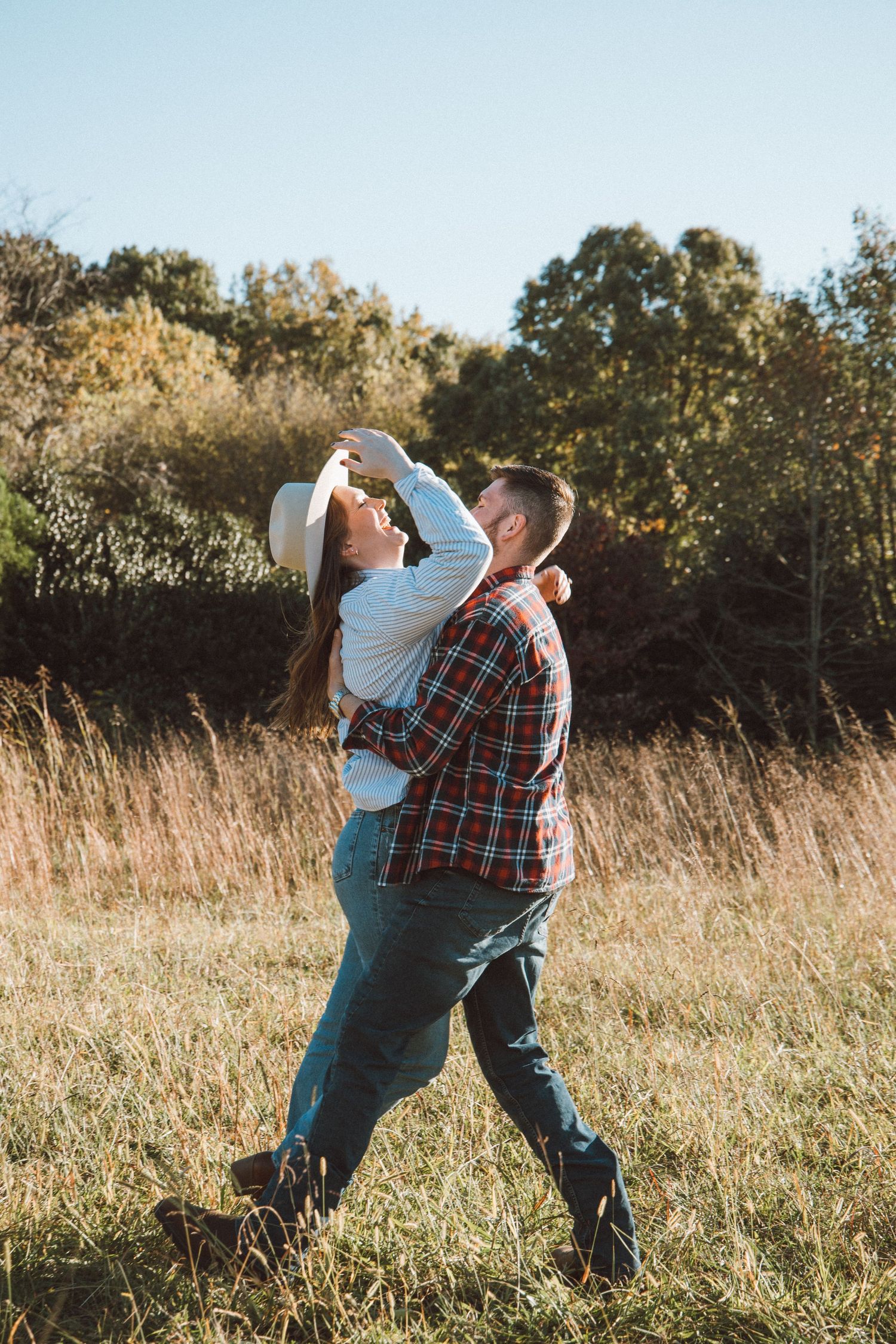 Couple dancing together in golden field during sunset with autumn trees in background.