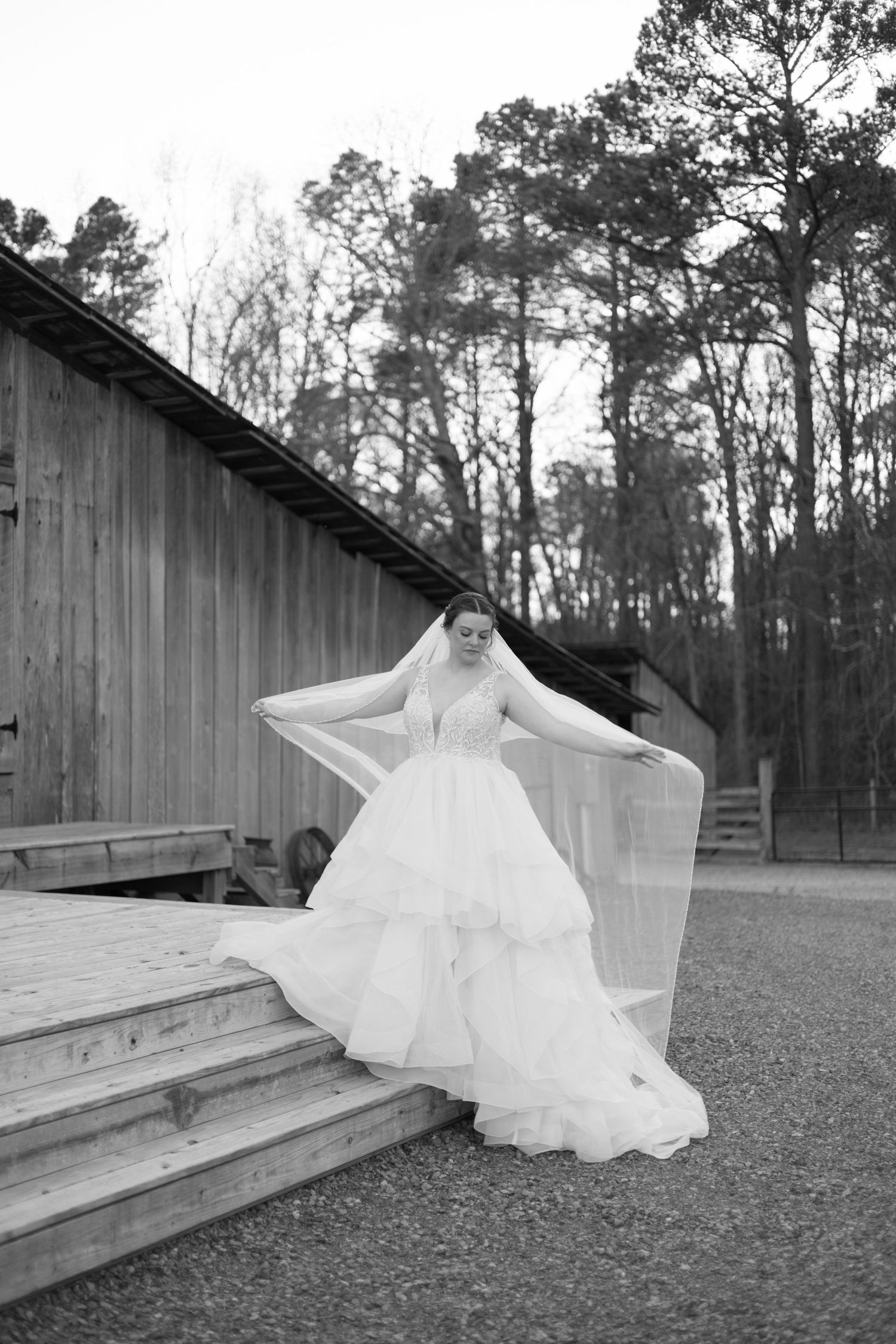 Black and white photo of a wedding dress on rustic wooden steps beside a barn with tall pine trees in background.