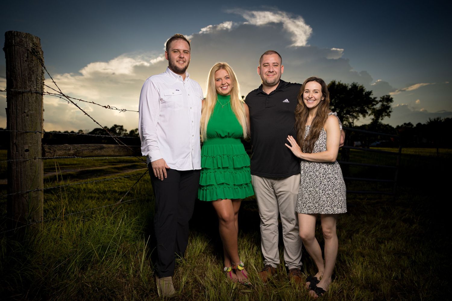 Family of four posing together in a green field at sunset near a rustic barbed wire fence.