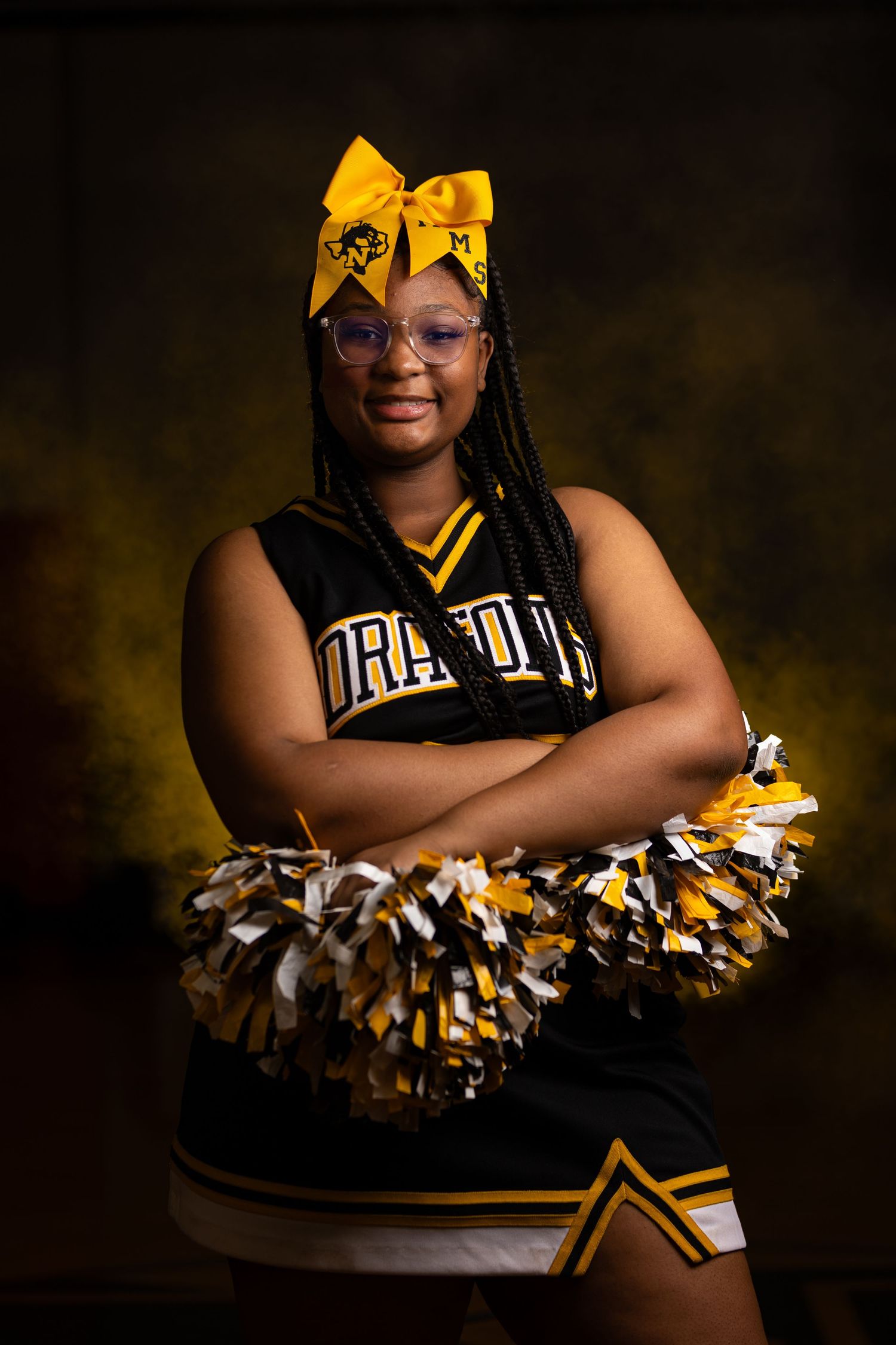 Cheerleader in black and gold uniform with yellow bow poses against dark background.