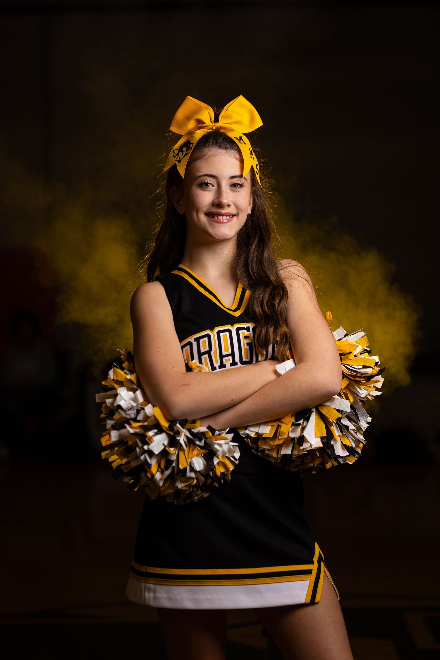 Cheerleader in Dragons uniform with yellow bow and pom poms poses against dramatic backdrop.