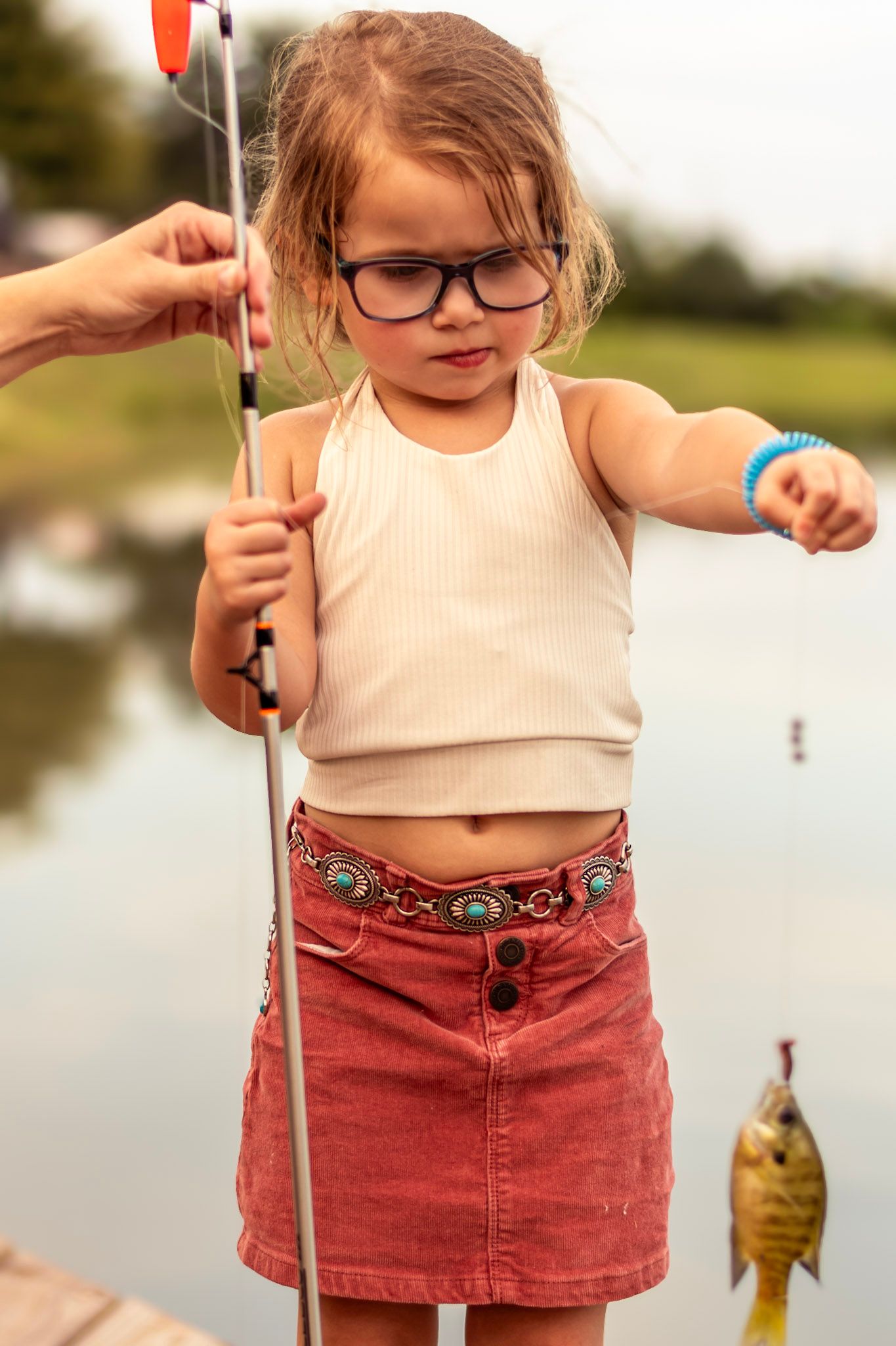 Young girl holding a fishing rod with proud smile, golden sunset light reflecting on the water.