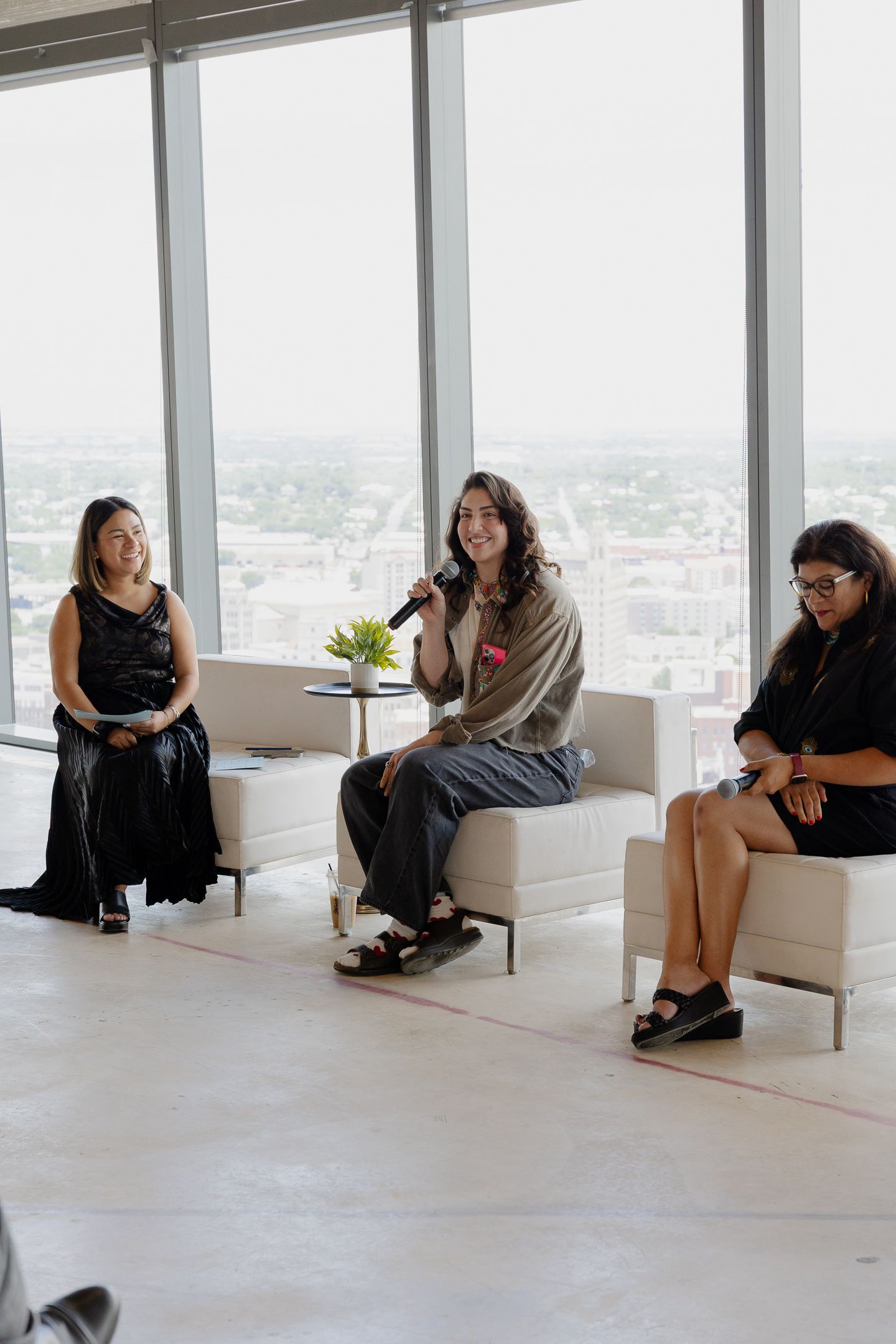A group of people sit spaced apart in white chairs along a bright modern room with floor-to-ceiling windows and city views.