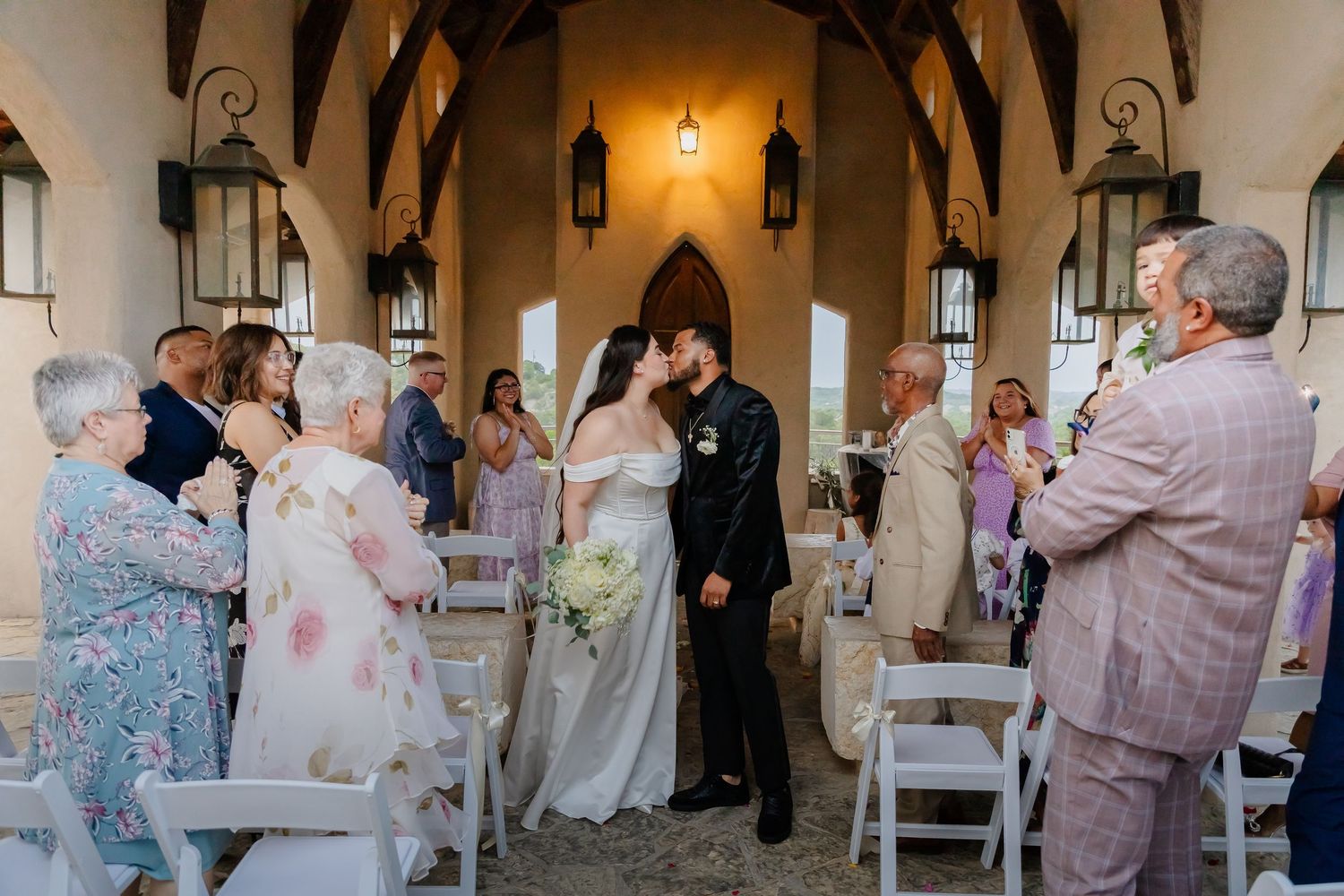 A romantic wedding ceremony takes place inside a rustic chapel with wooden beams and hanging lanterns.