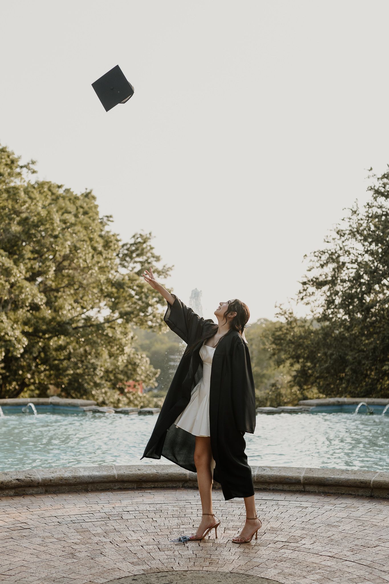 Graduate in black academic regalia tossing cap by fountain.