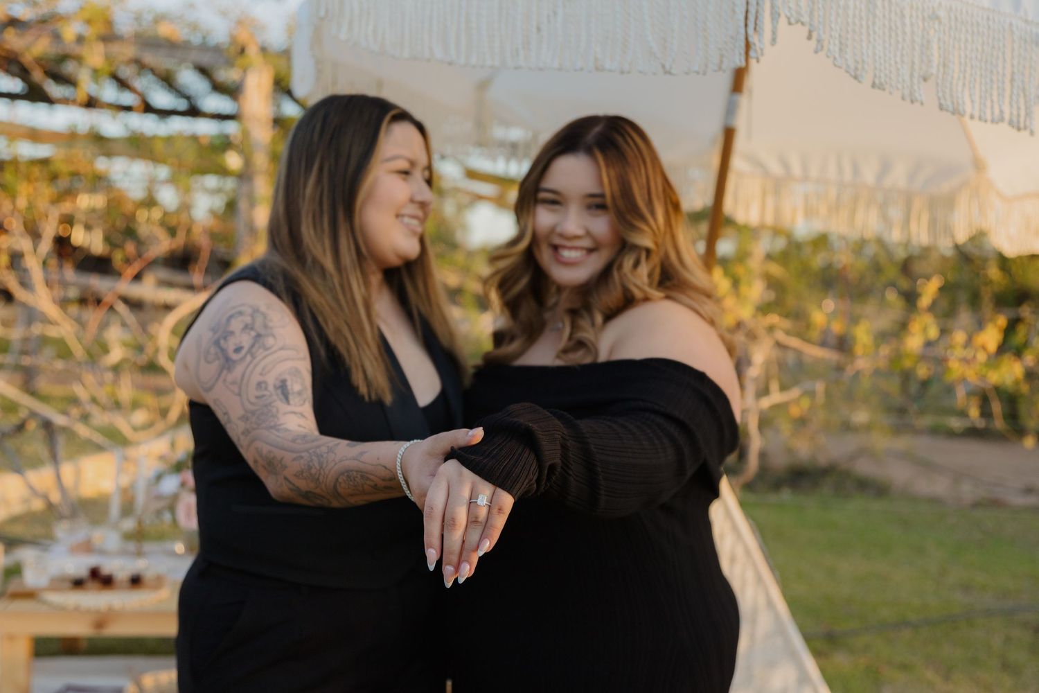 Two people in black outfits share a tender moment outdoors at sunset with string lights in the background.