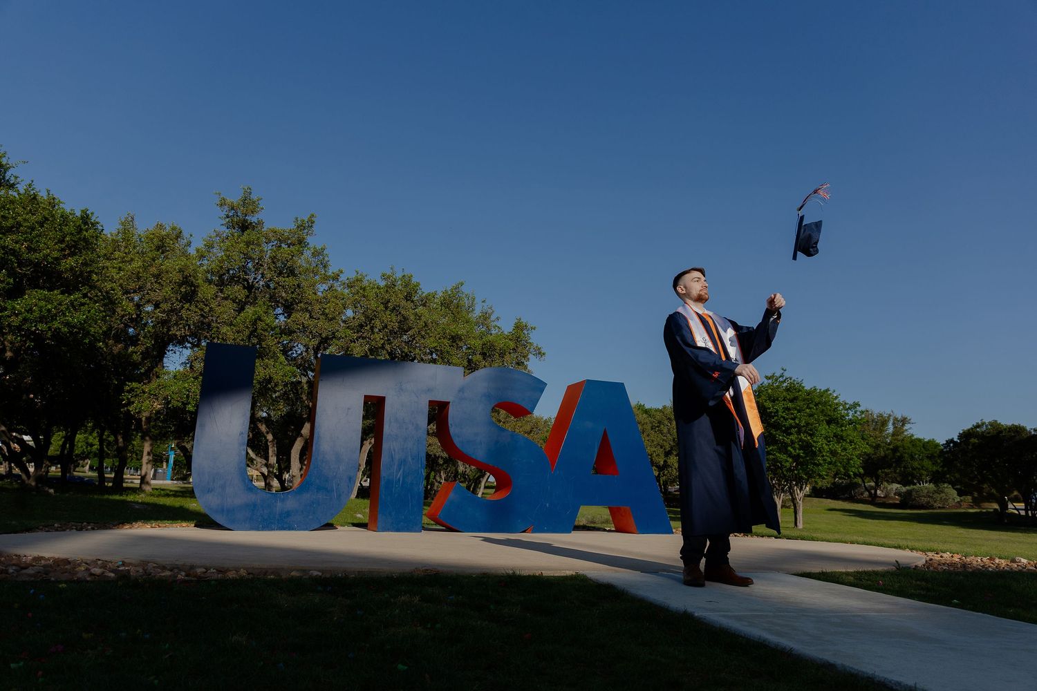 Graduate celebrates by UTSA sign while tossing cap against blue sky.