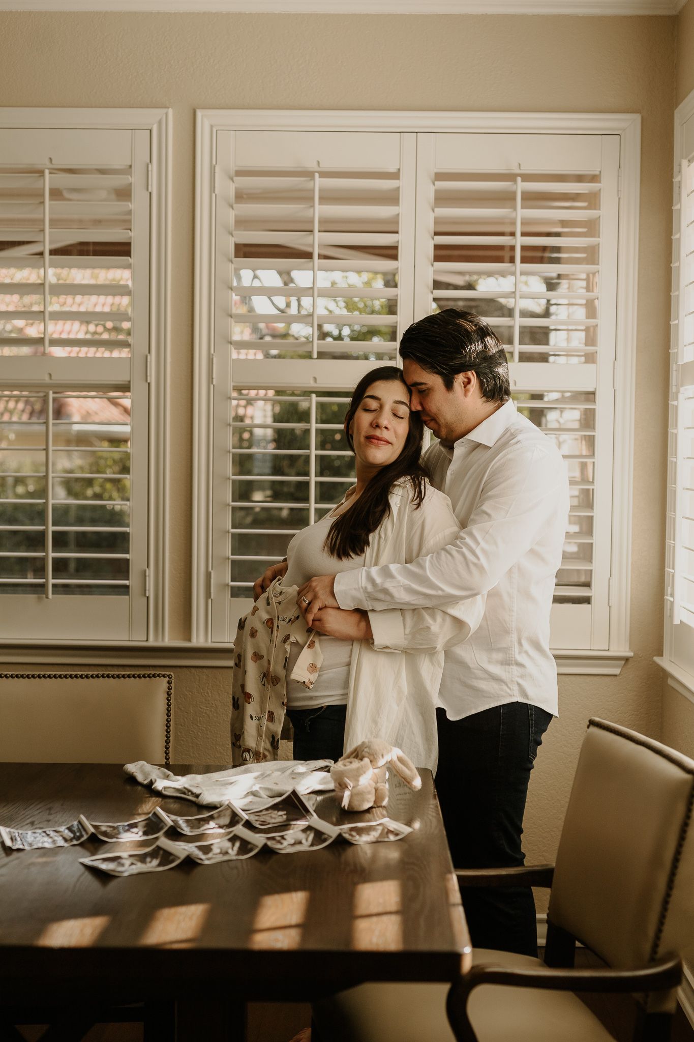 Intimate moment between expecting couple standing by window with ultrasound photos on table.
