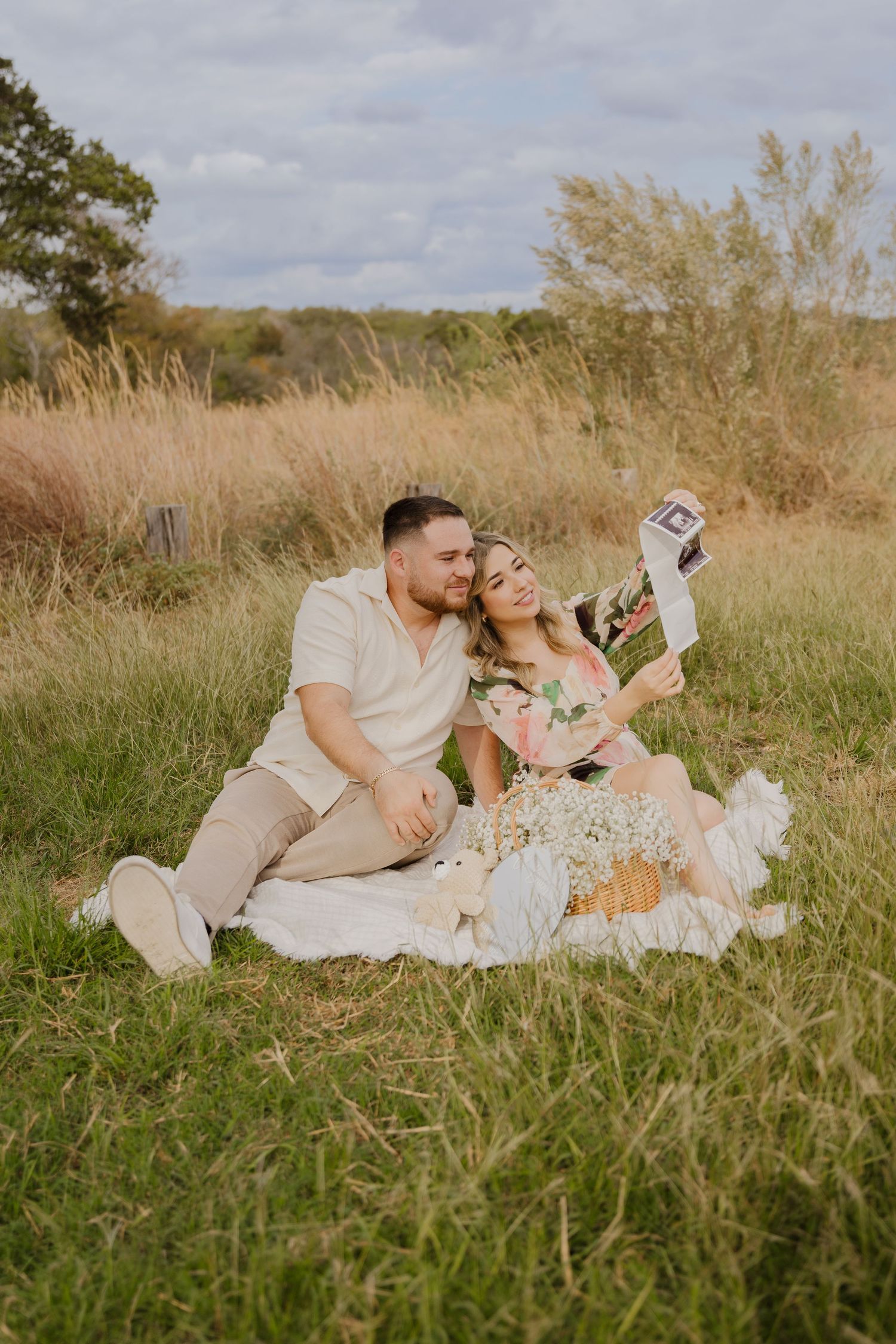 Couple sitting on blanket in field sharing pregnancy announcement photos.