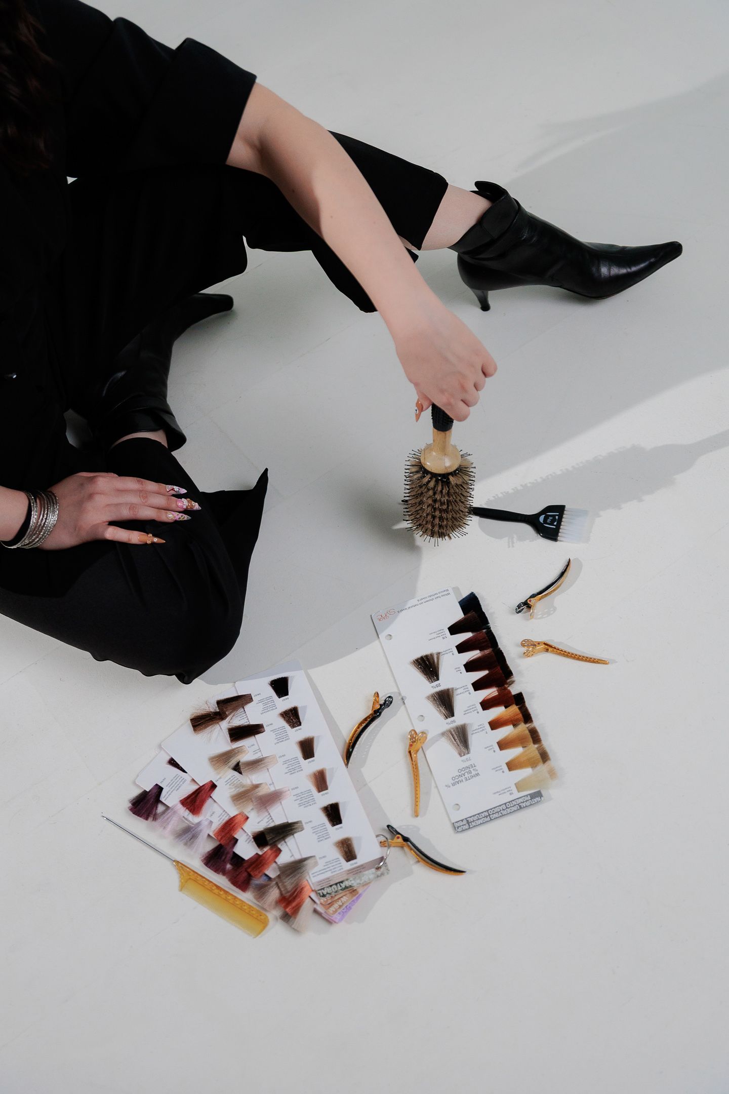 Person in black outfit sitting on floor with hair color swatches, round brush, and styling tools spread out.