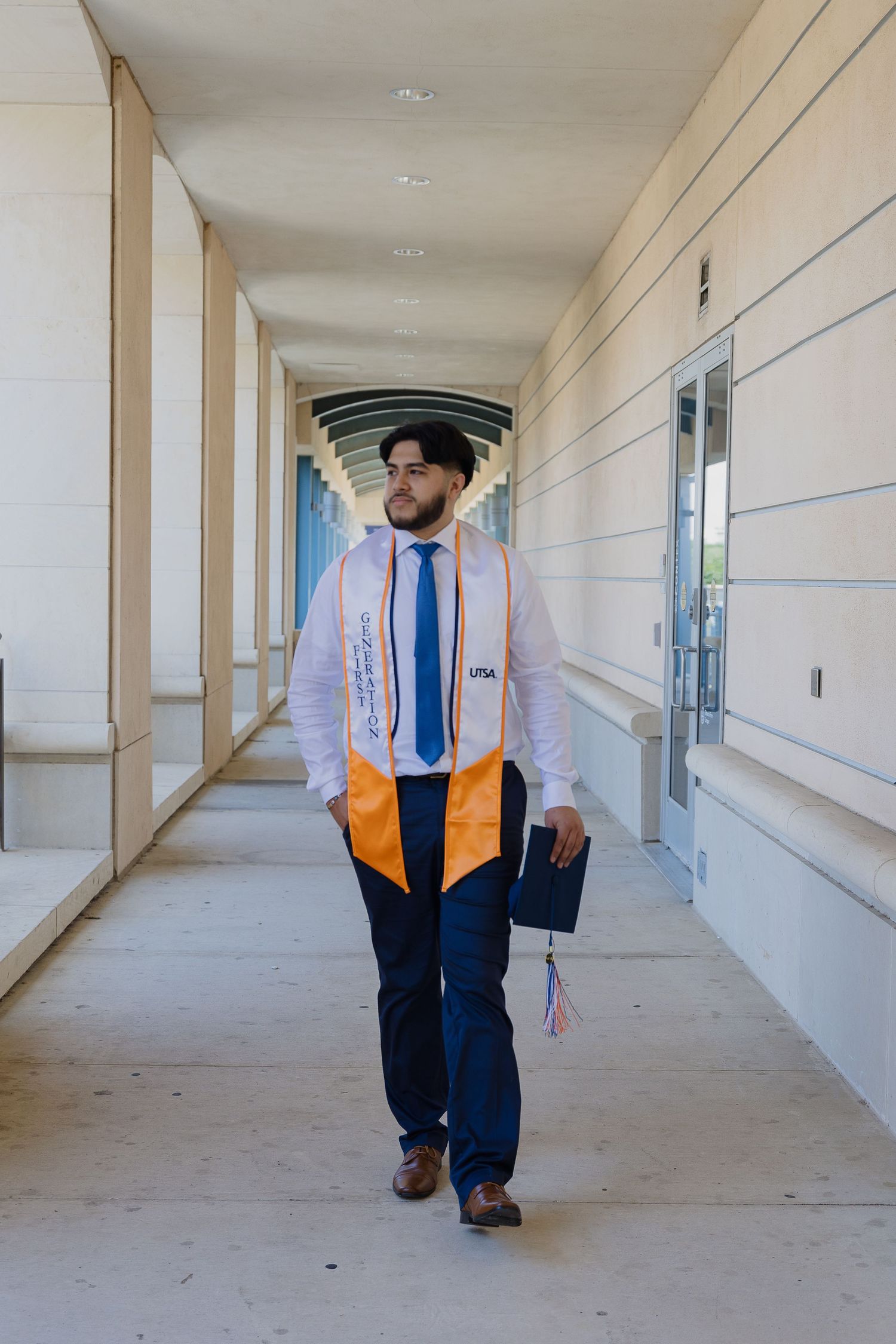 Graduate student wearing graduation regalia walks through a covered outdoor hallway at a university campus.
