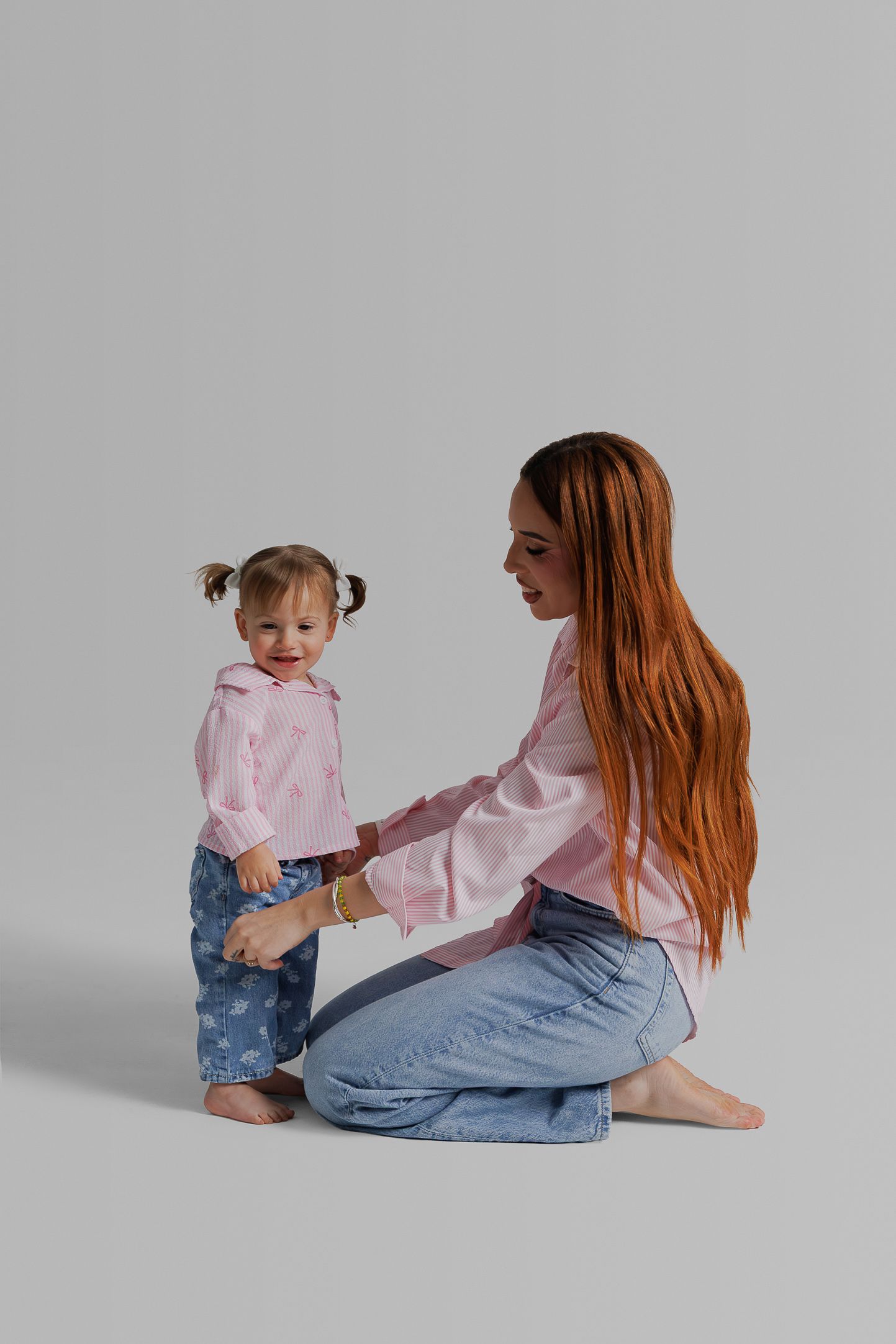 Mother and toddler in matching pink shirts and jeans pose together against grey background in casual studio setting.