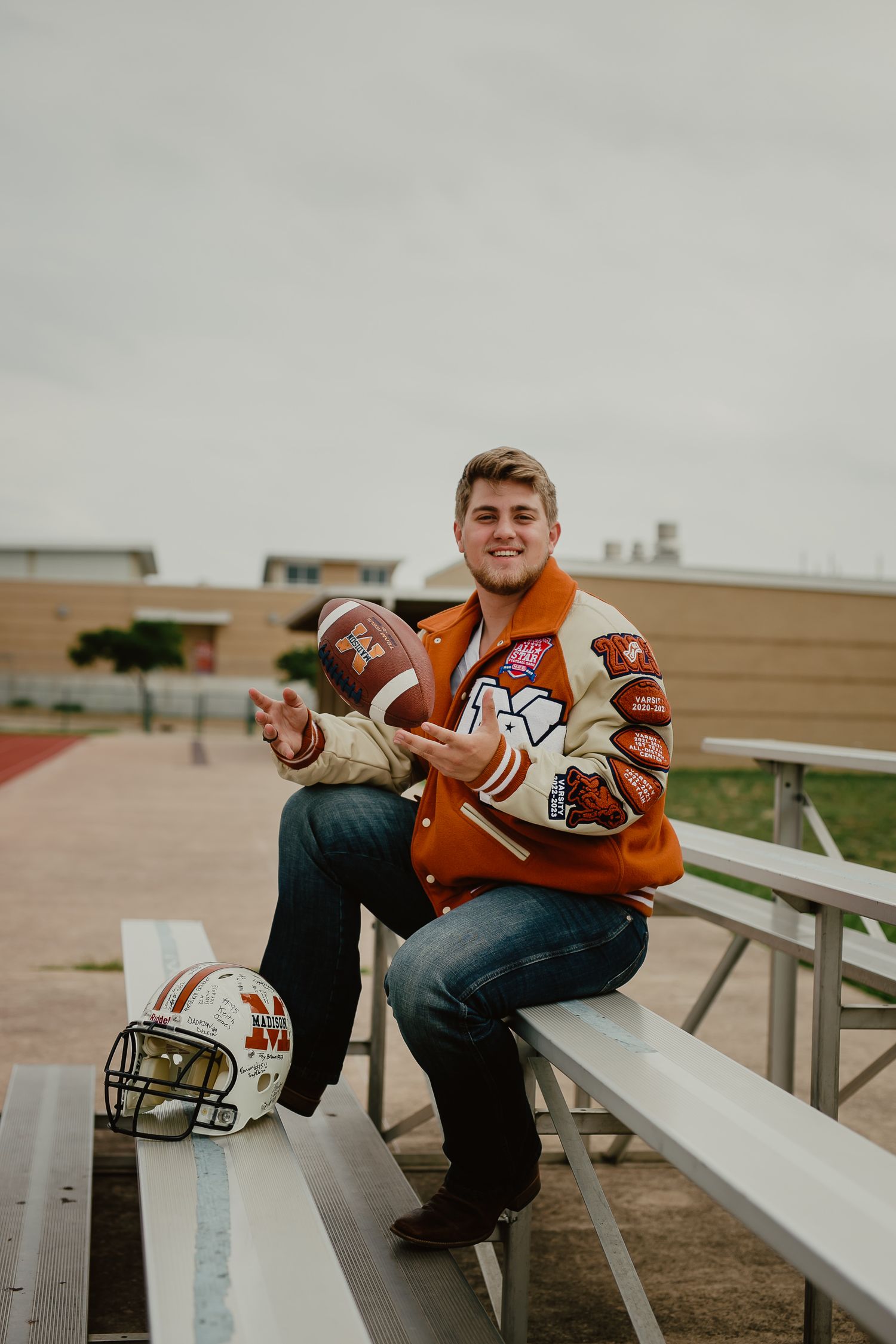 Two students in football gear pose playfully on bleachers at a high school stadium.